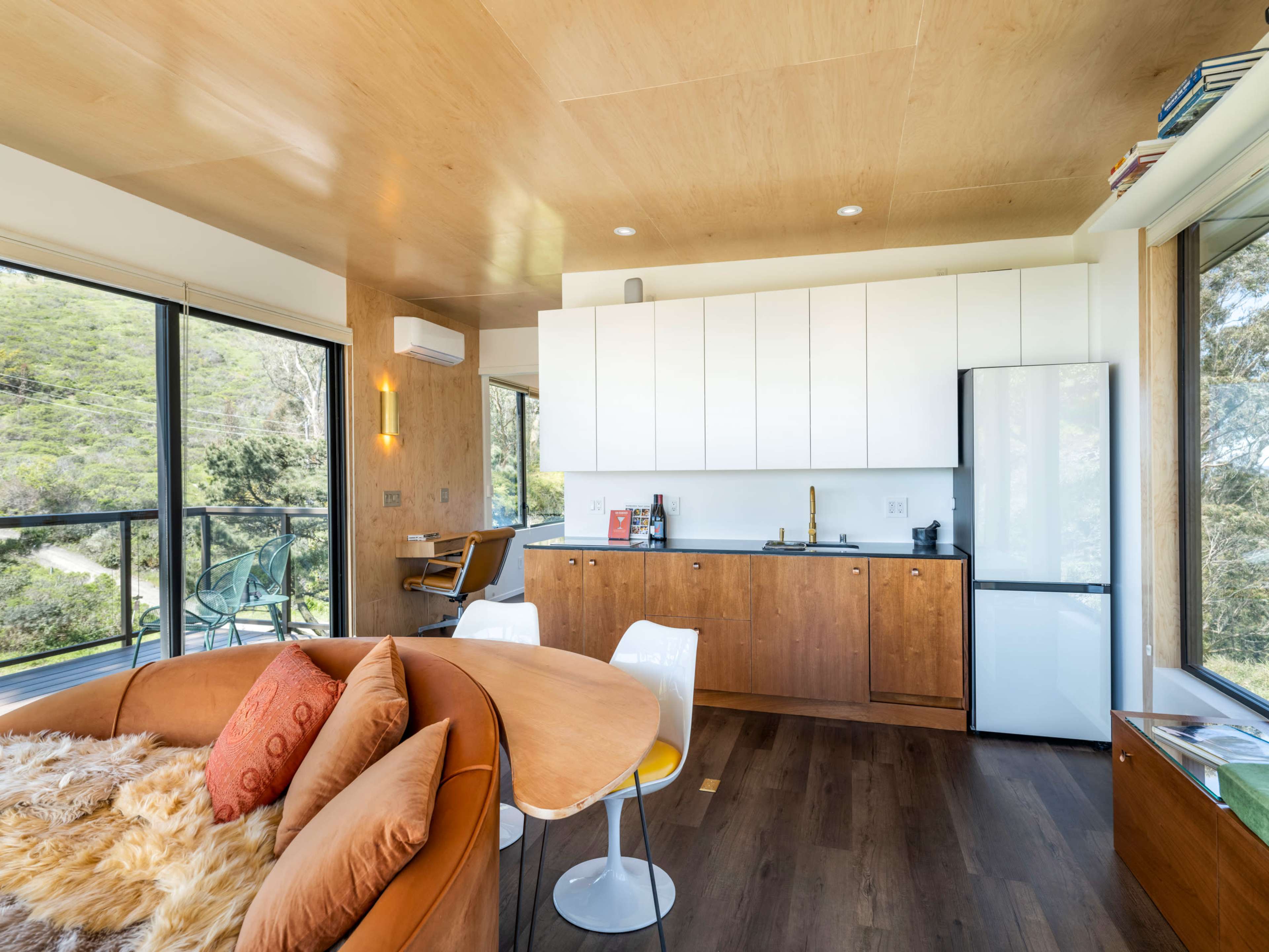The image shows a modern, open-concept living space featuring a kitchen with white cabinets, a wooden dining table, and large windows overlooking greenery.