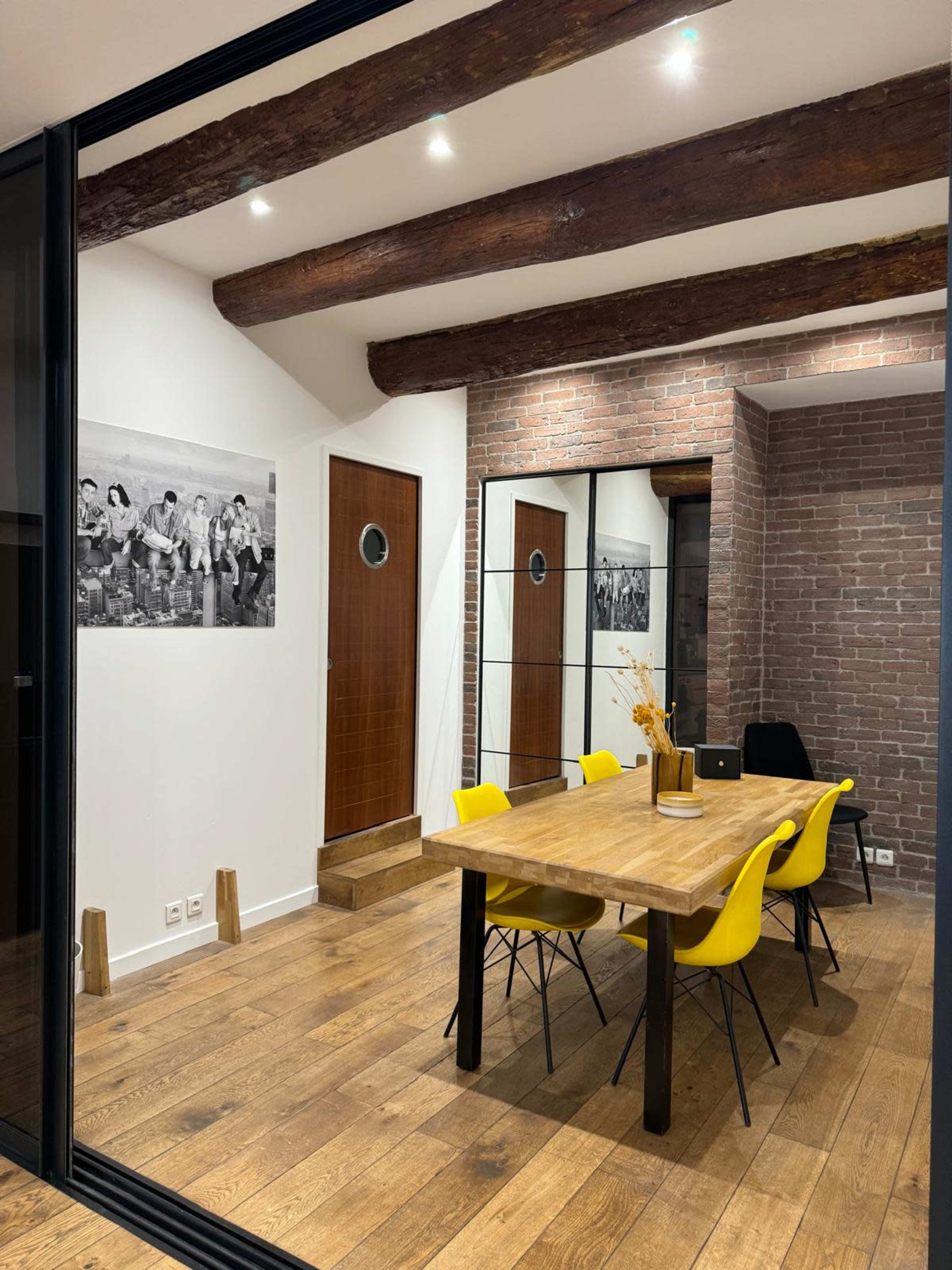 A modern conference room features a wooden table surrounded by yellow chairs, with exposed brick walls and large windows.