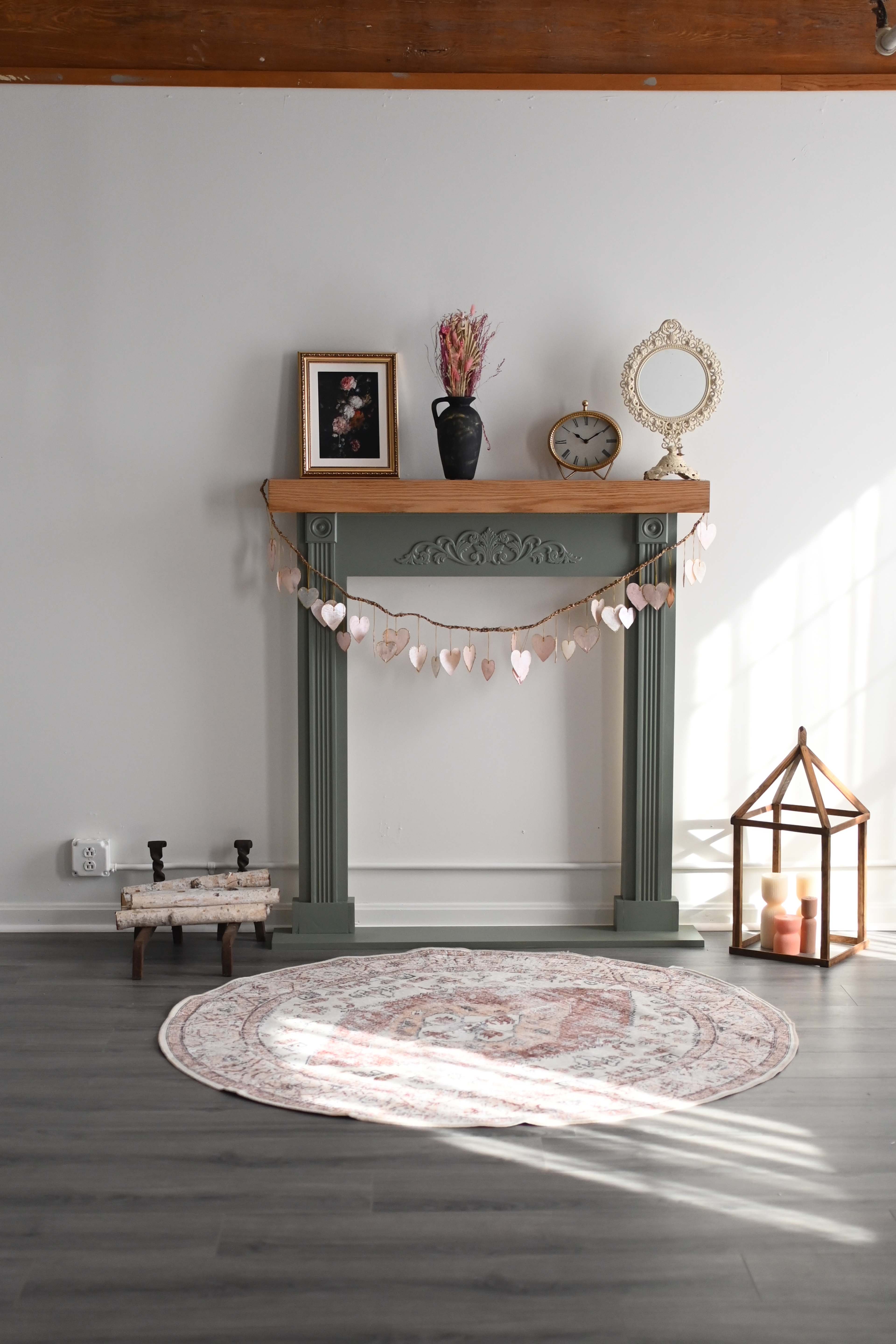 A decorative fireplace mantel is adorned with a vase, a clock, and a garland of heart shapes, accompanied by a lantern and a round rug on the floor.