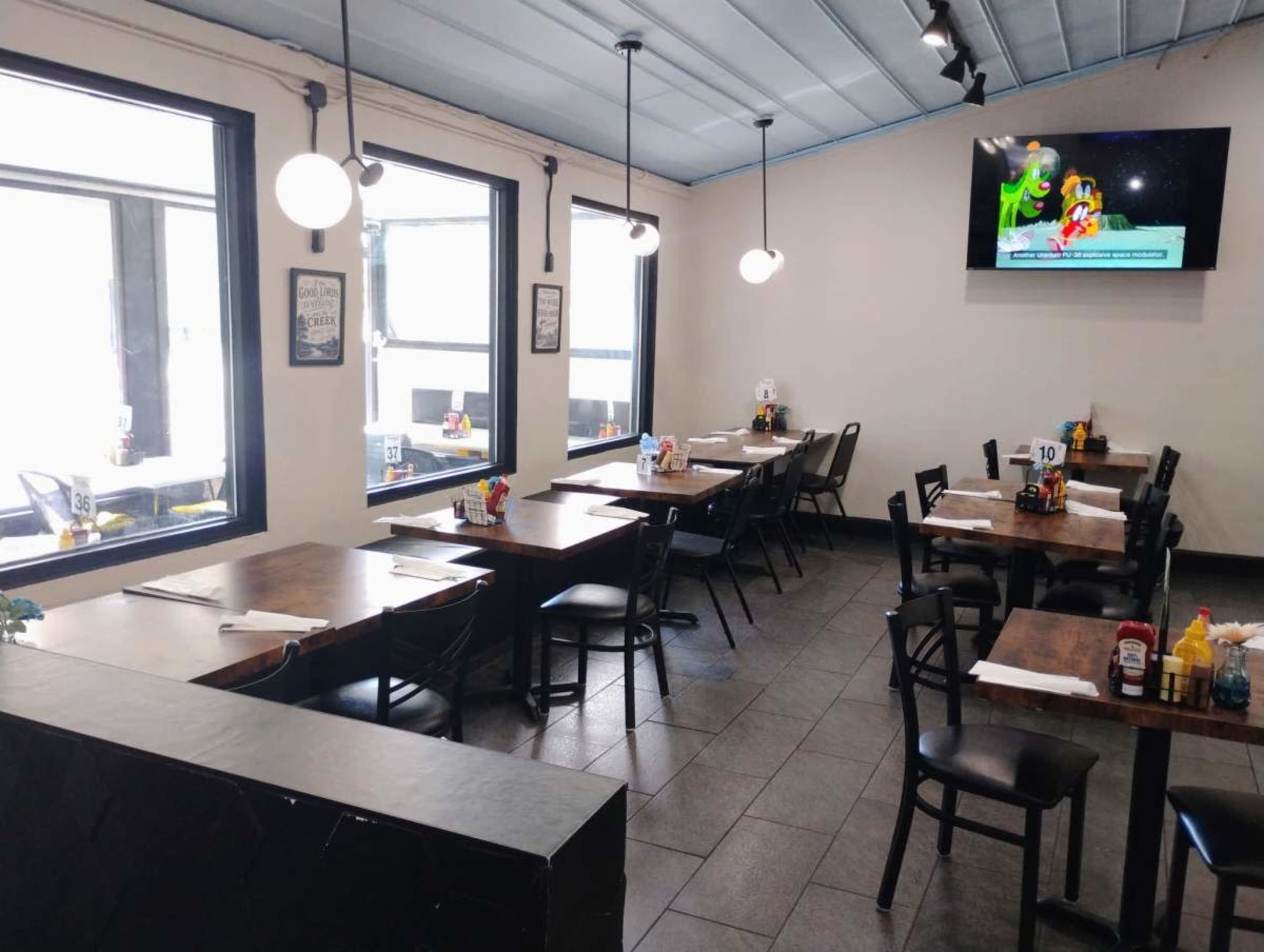 The image shows a modern restaurant interior with tables arranged beside large windows and a television mounted on the wall displaying a cartoon.