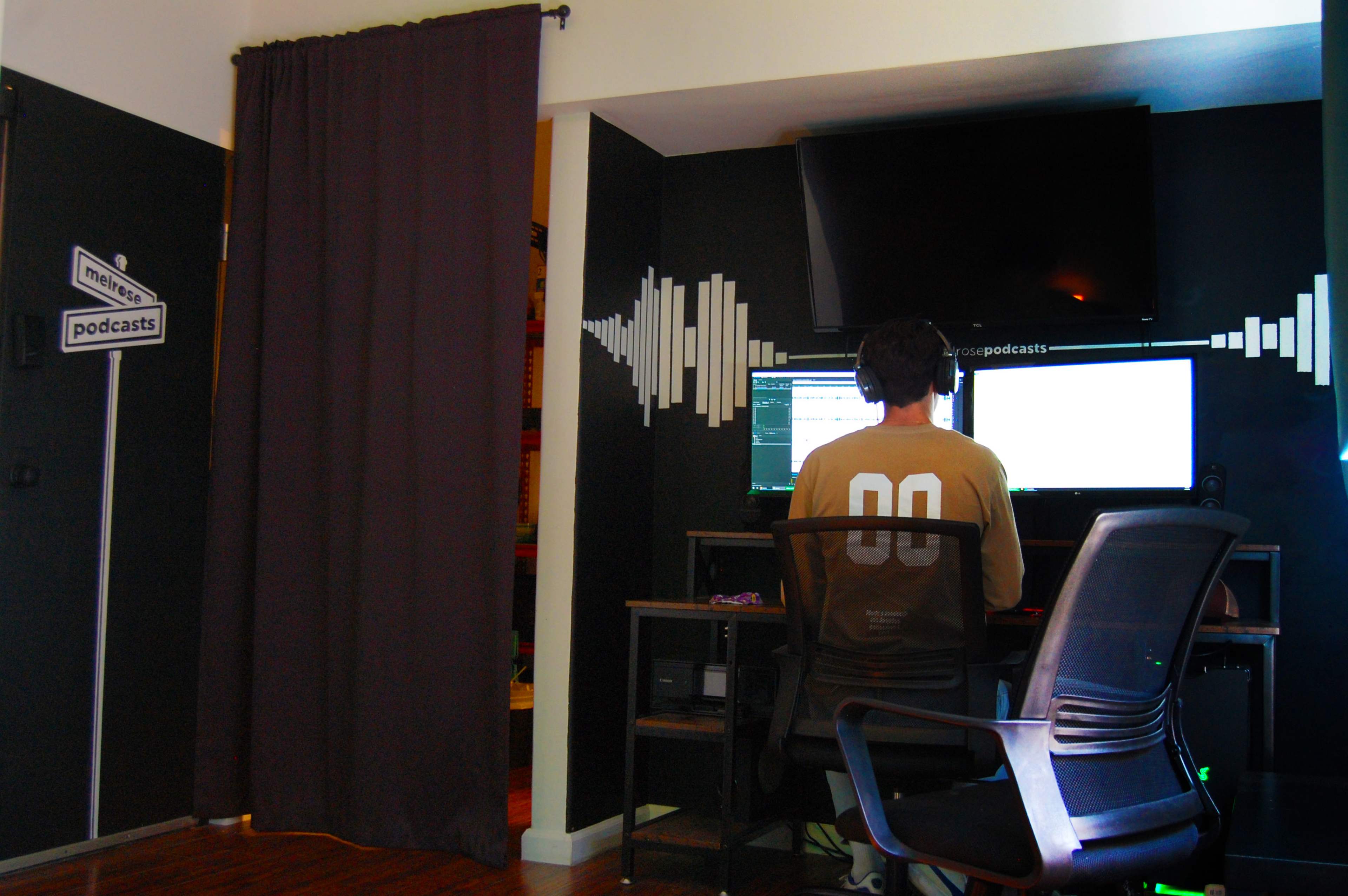A person sits at a desk with two monitors in a room decorated for podcasting, featuring a dark curtain and soundwave graphics on the wall.
