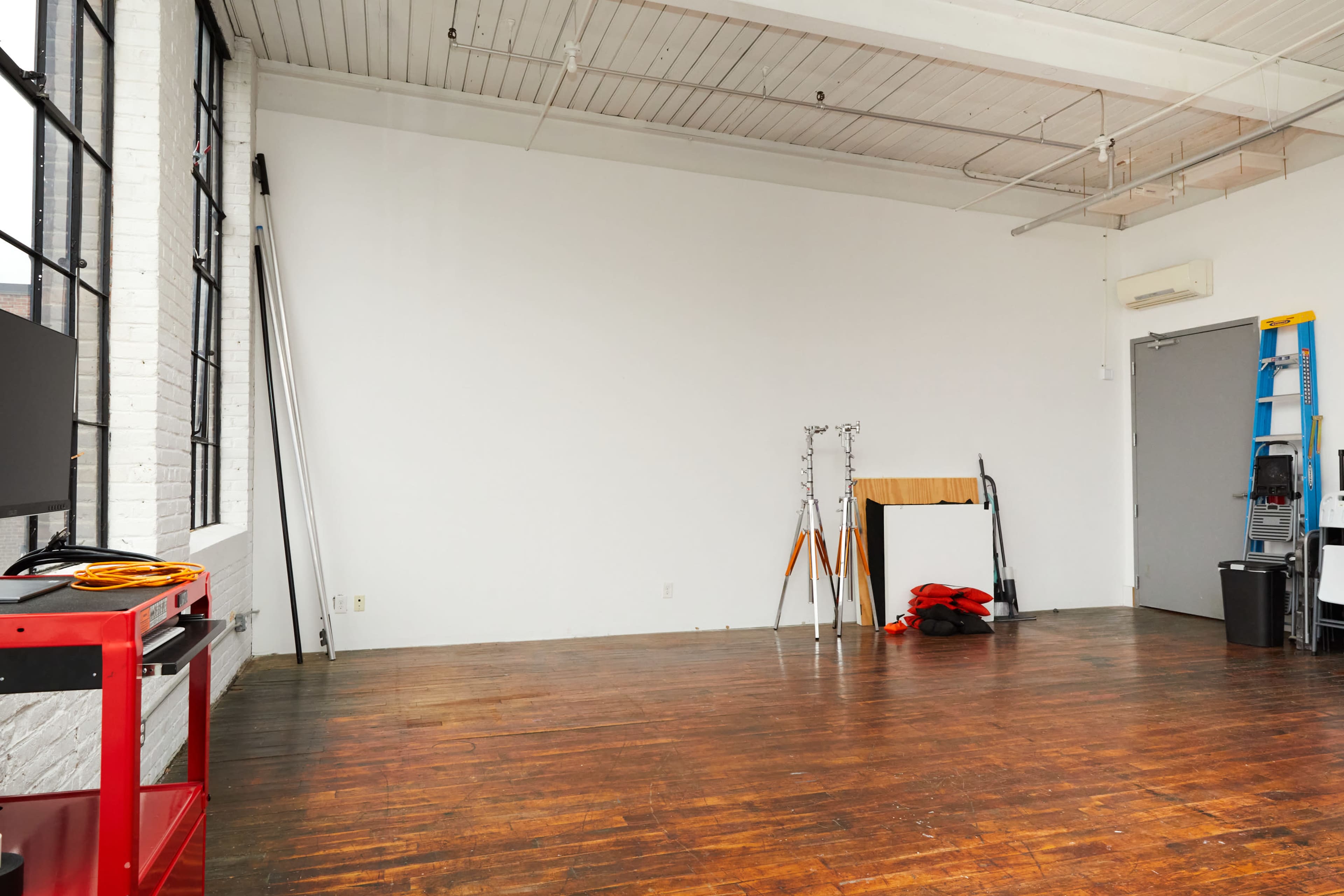 A spacious studio with wooden flooring, large windows, and minimal furnishings, including a ladder, a small table, and various equipment against the walls.