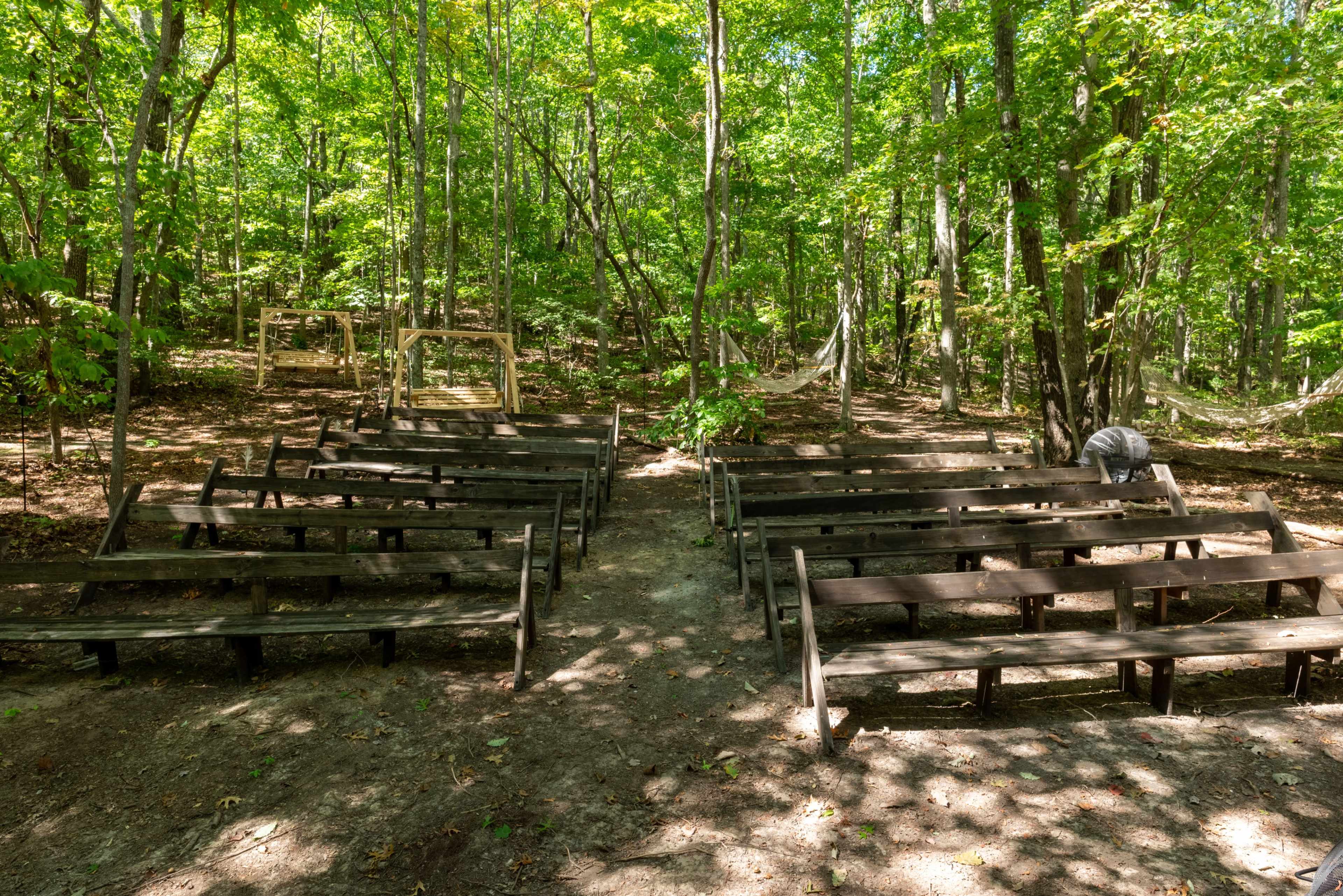 A clearing in the woods features several rows of wooden benches arranged for an outdoor gathering, with a swingset visible in the background.