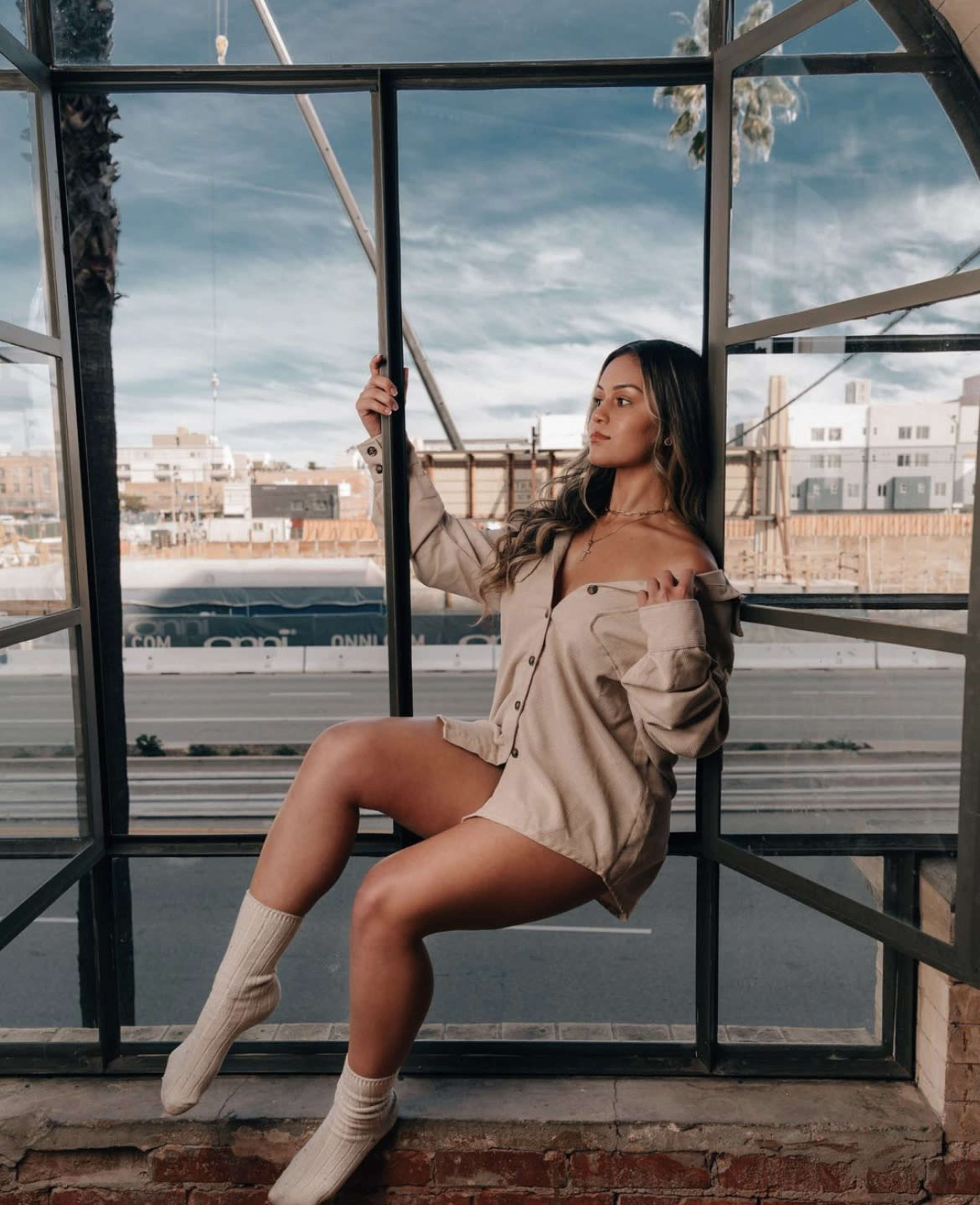 A person sits on a window ledge, dressed in an oversized shirt and knee-high socks, with a cityscape visible through the window behind them.