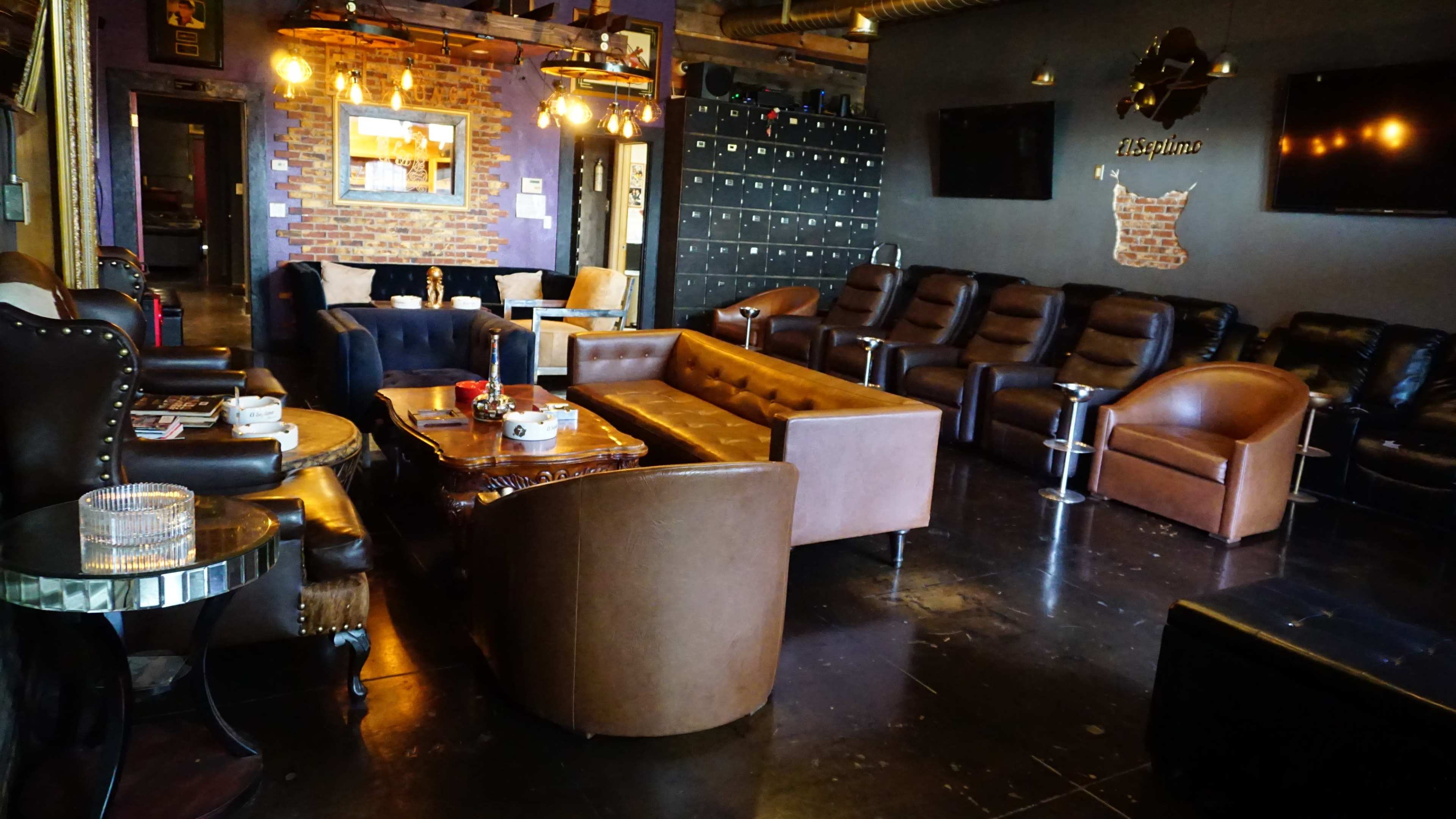 The image shows a modern lounge space with various seating options, including leather couches and recliners, and decor featuring exposed brick walls and industrial lighting.