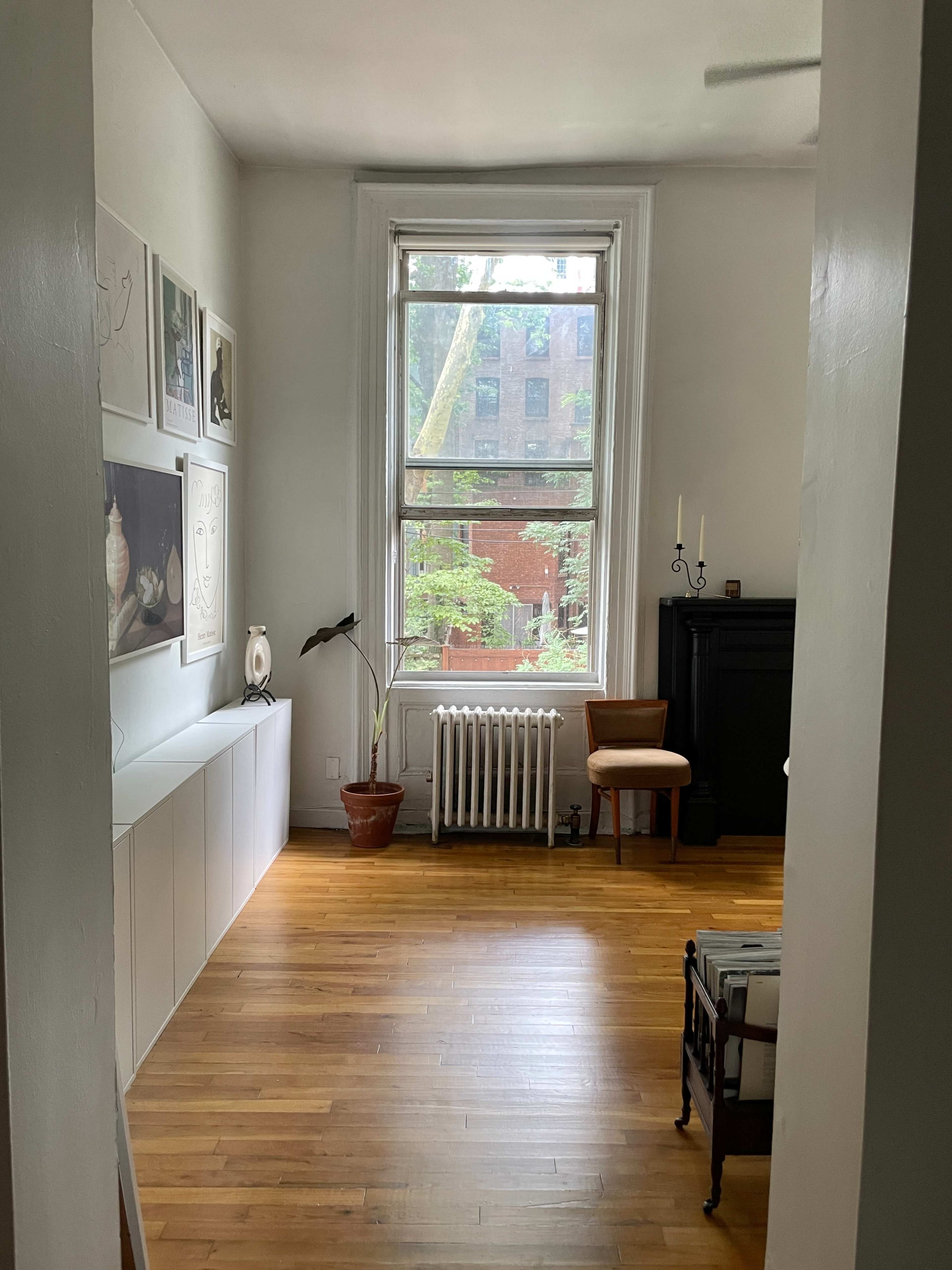A spacious room with wooden floors, a large window overlooking greenery, and minimal furniture including a chair and a radiator.
