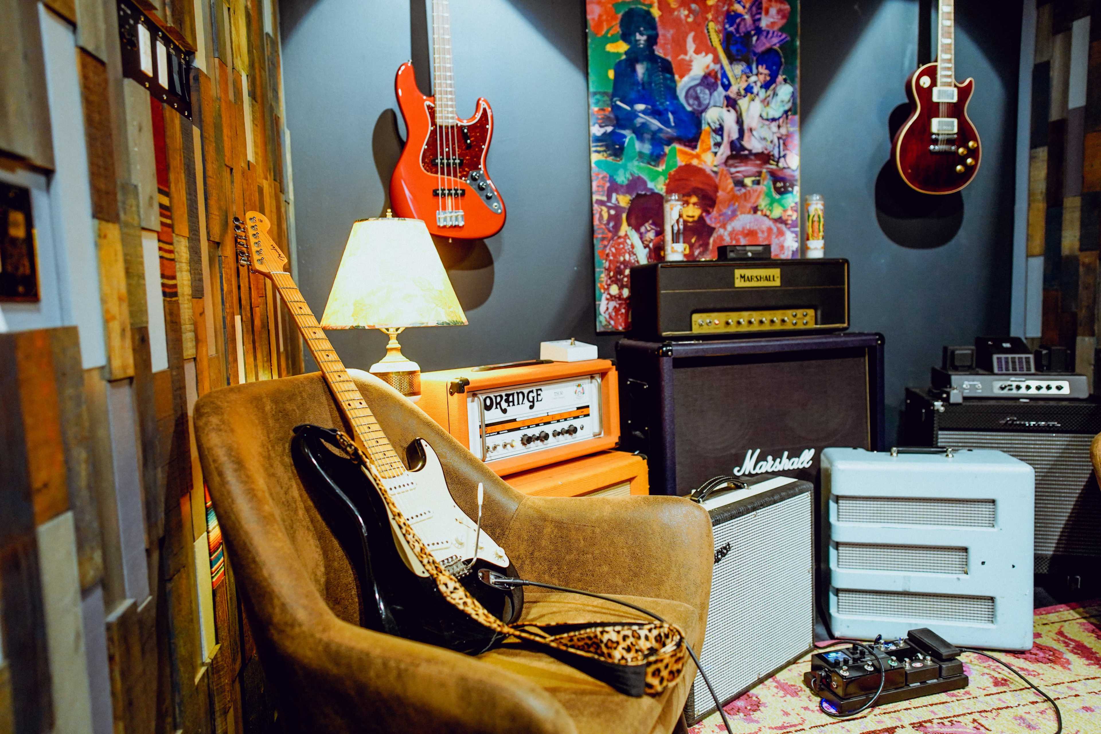The image shows a cozy music room featuring various guitars mounted on the wall, amplifiers and a lamp beside a guitar resting on an armchair.