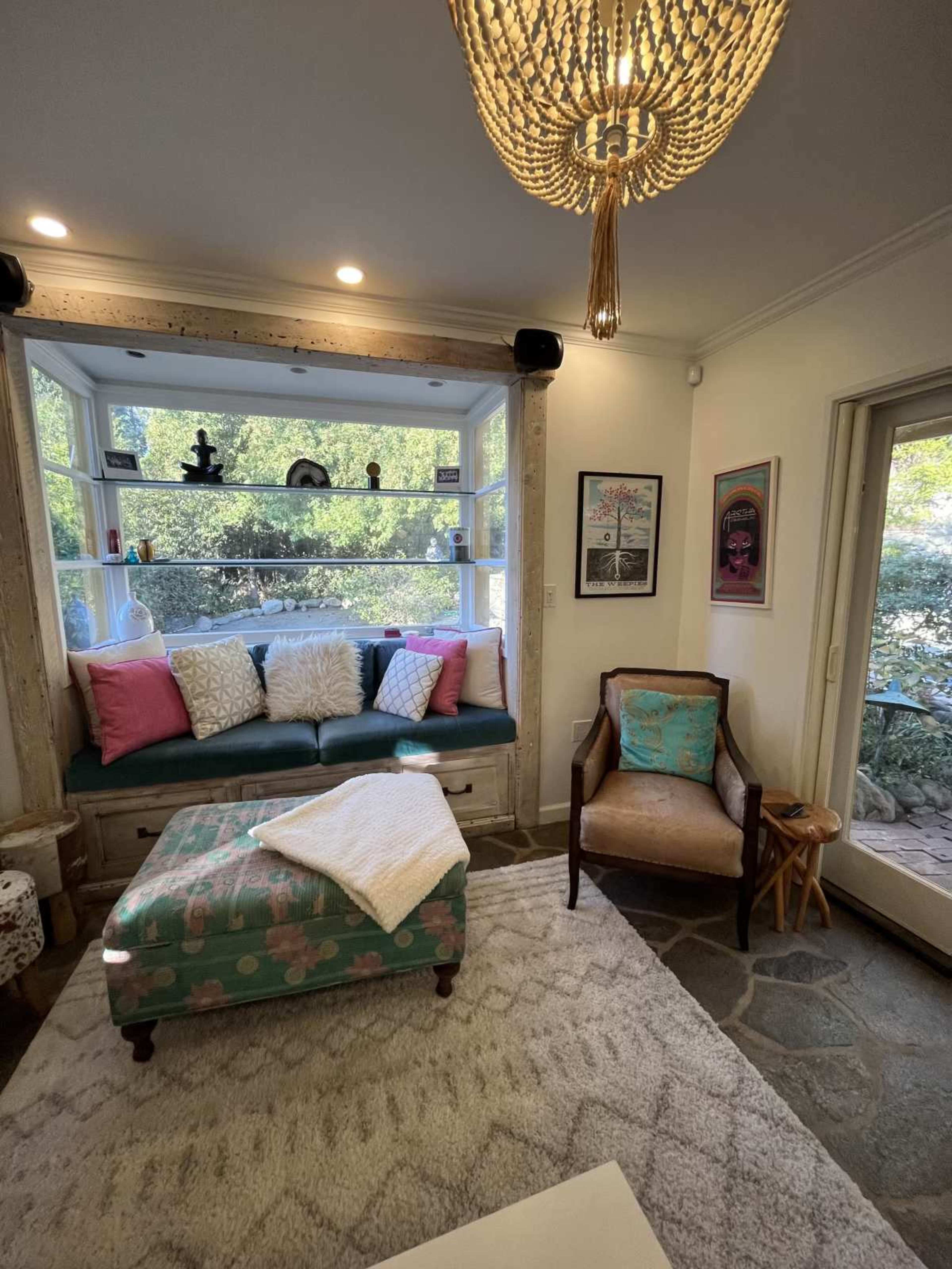 The image shows a cozy living room with a built-in seating area, colorful cushions, a coffee table, and a stone floor, illuminated by a chandelier.