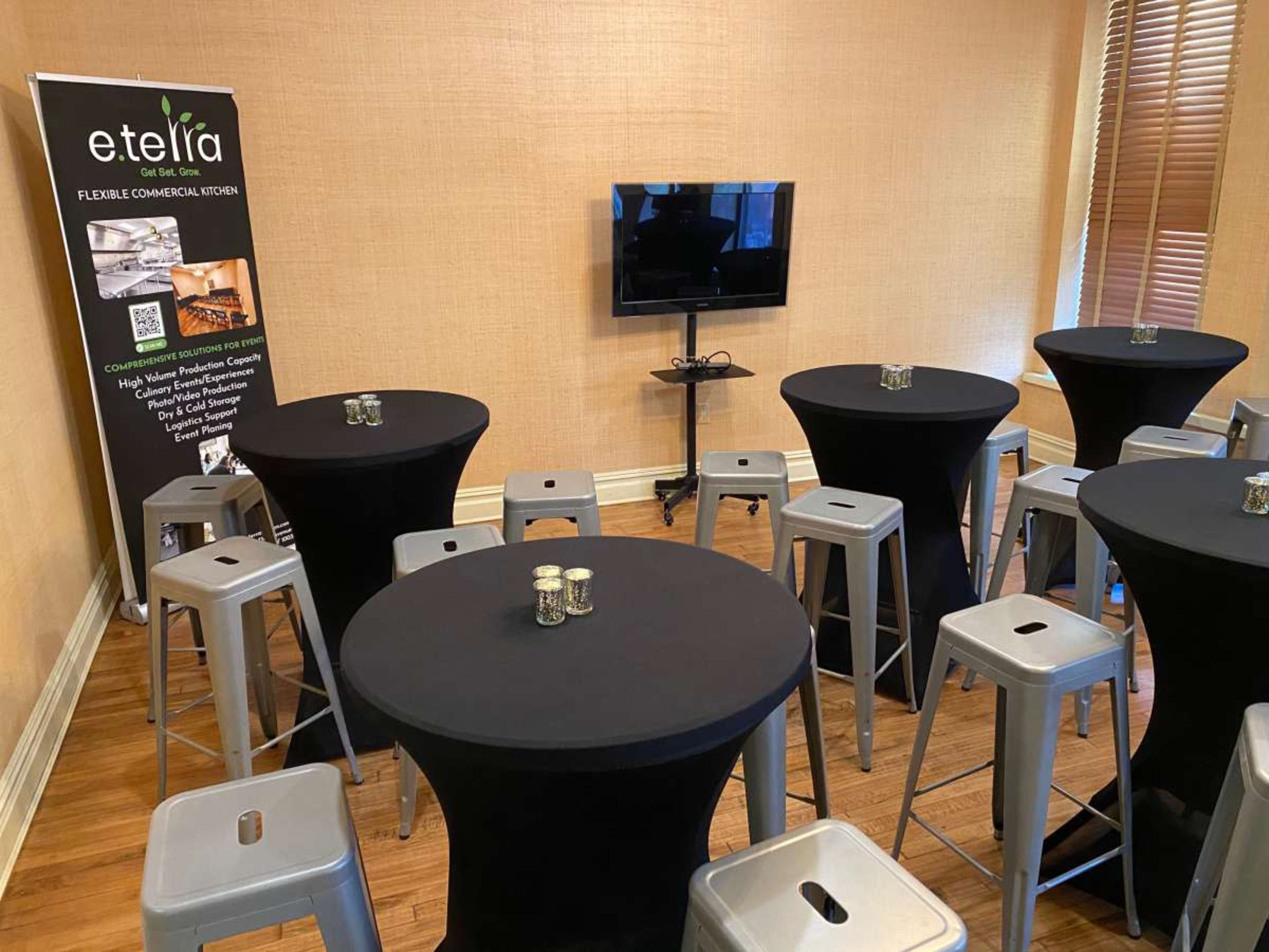 The image shows a room set up for an event, featuring several black tables with high stools, a television mounted on the wall, and a promotional banner for e.tiara.