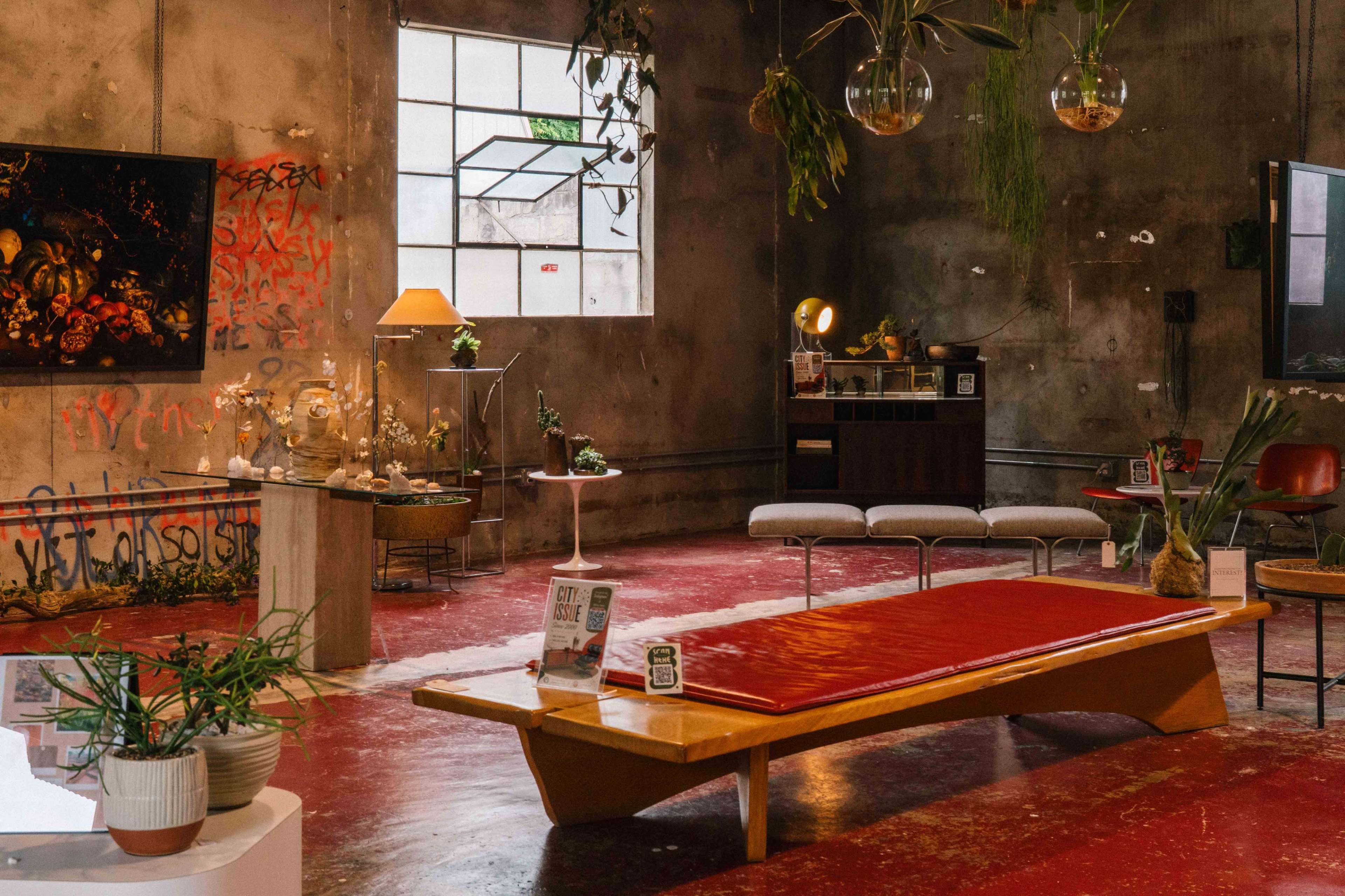 The image shows an industrial-style interior featuring a red floor, minimalist furniture, plants, and various decorative objects against a backdrop of graffiti on the walls.
