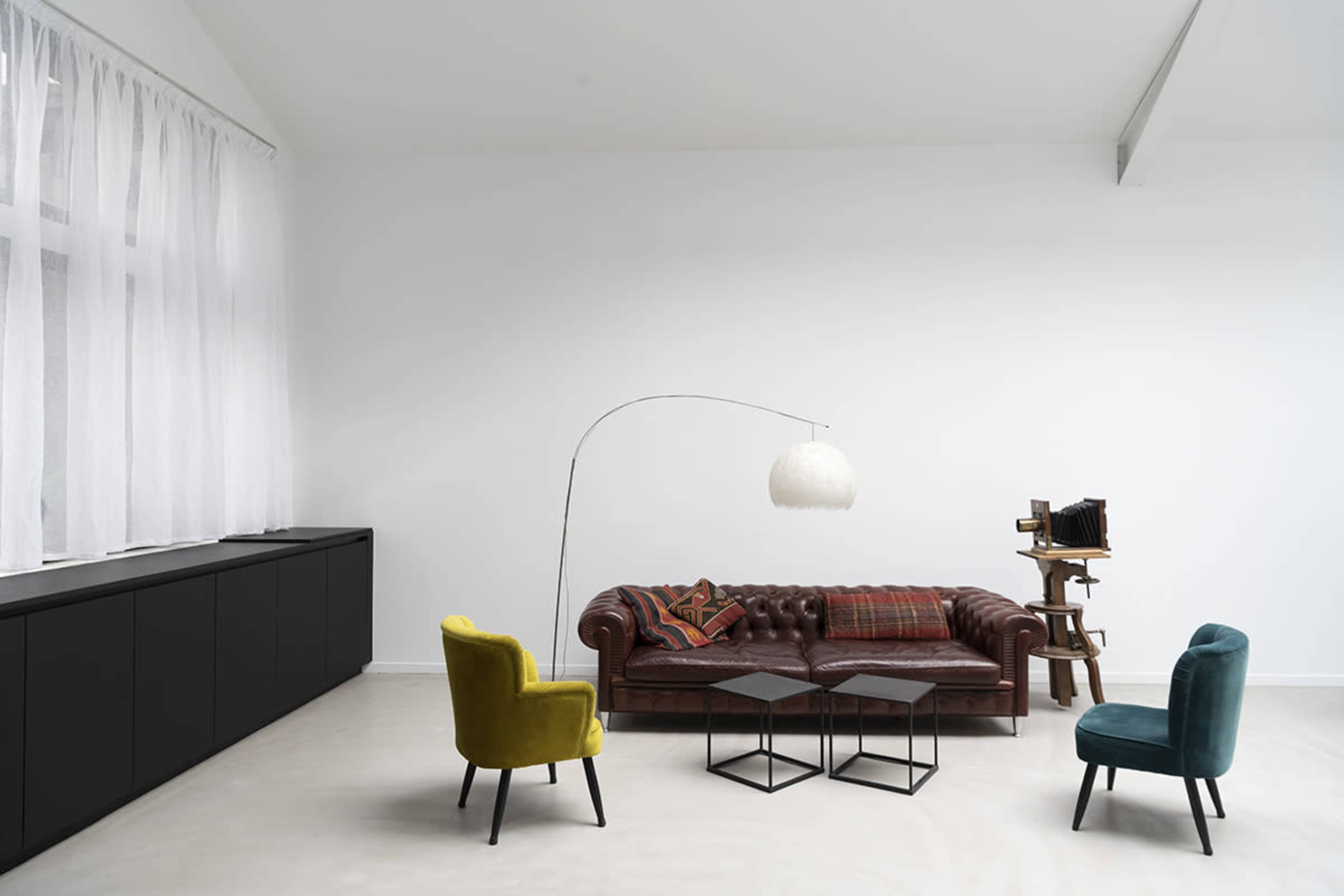 A living room with a brown leather chesterfield sofa, two colorful chairs, a floor lamp, and a wooden shelf against a white wall.