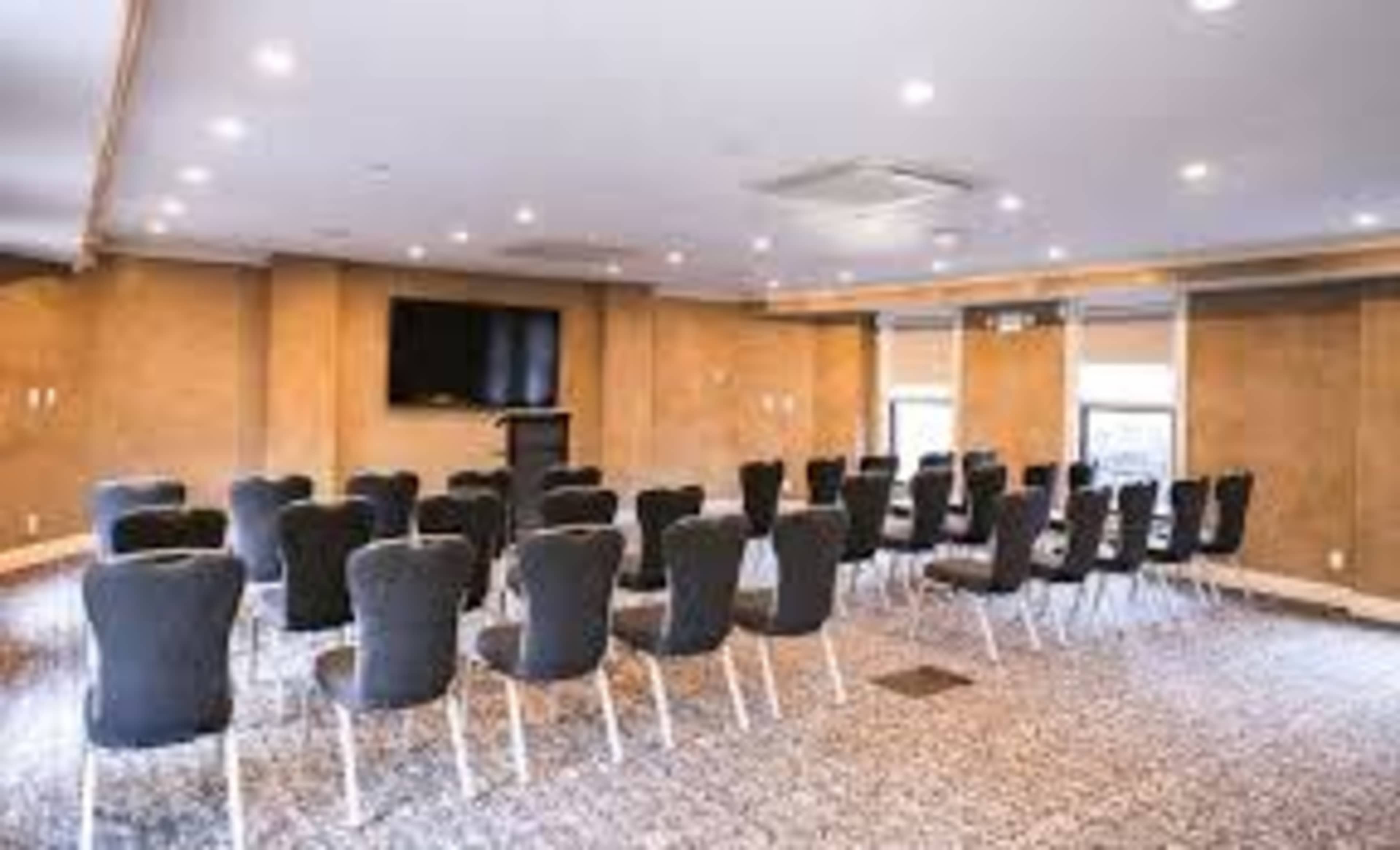 A conference room is arranged with rows of chairs facing a presentation area and a screen on the wall.