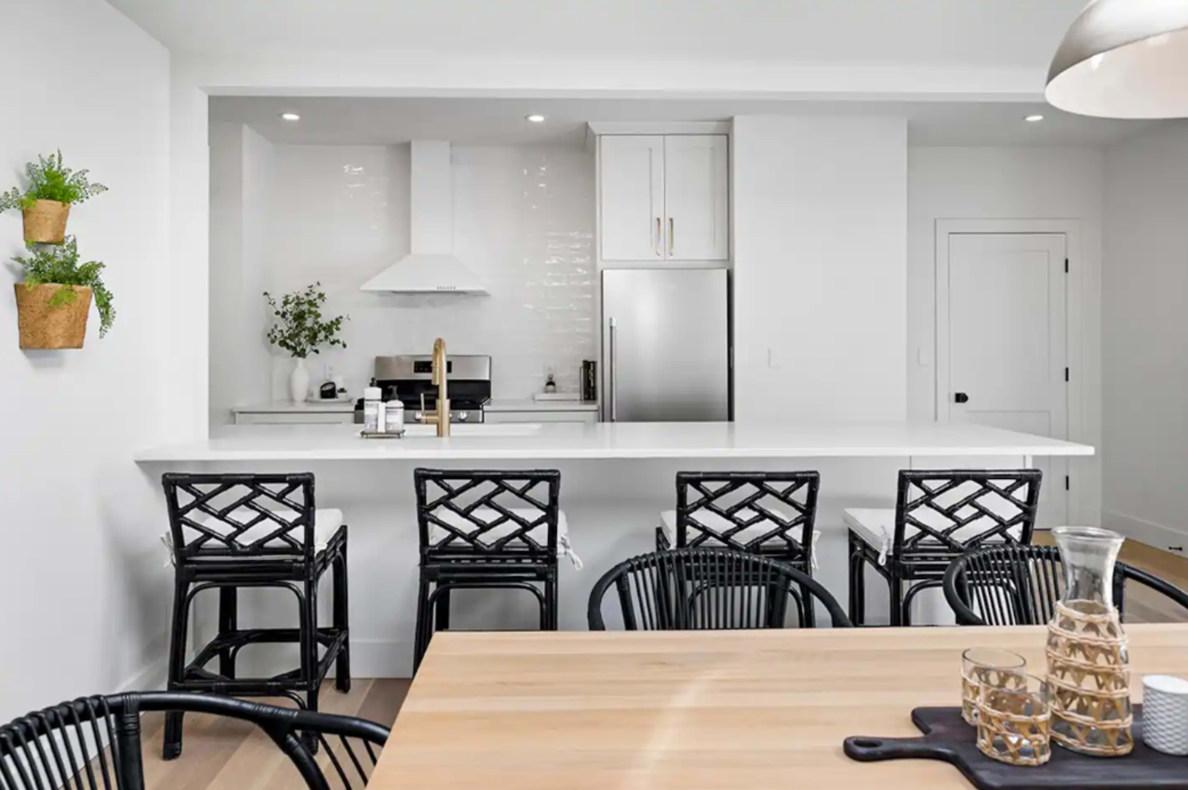 A modern kitchen features a white countertop island with four black bar stools, complemented by stainless steel appliances and minimal decor.