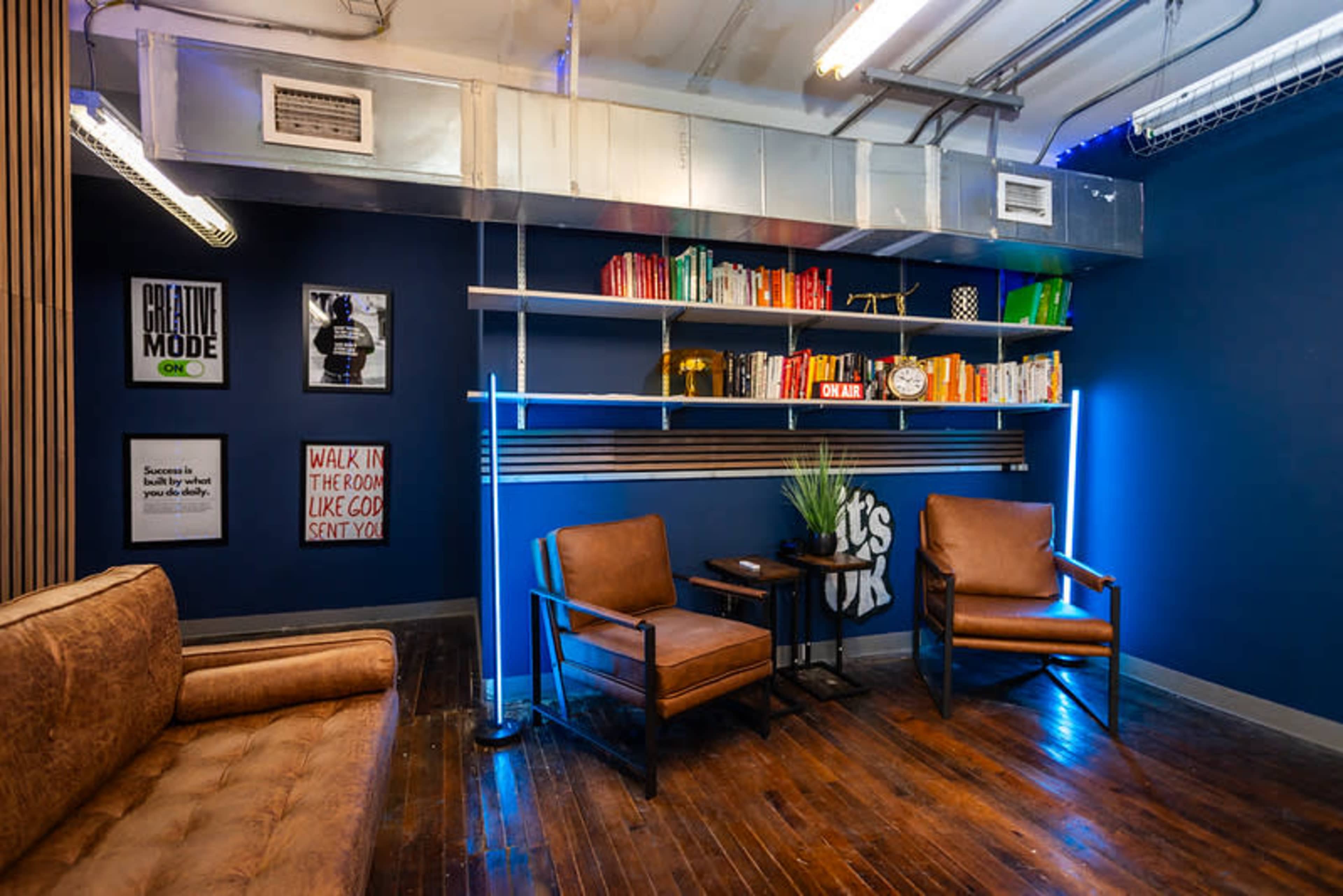 The image depicts a modern lounge area featuring a brown couch and two leather chairs, with a wall shelf displaying colorful books and framed motivational posters.