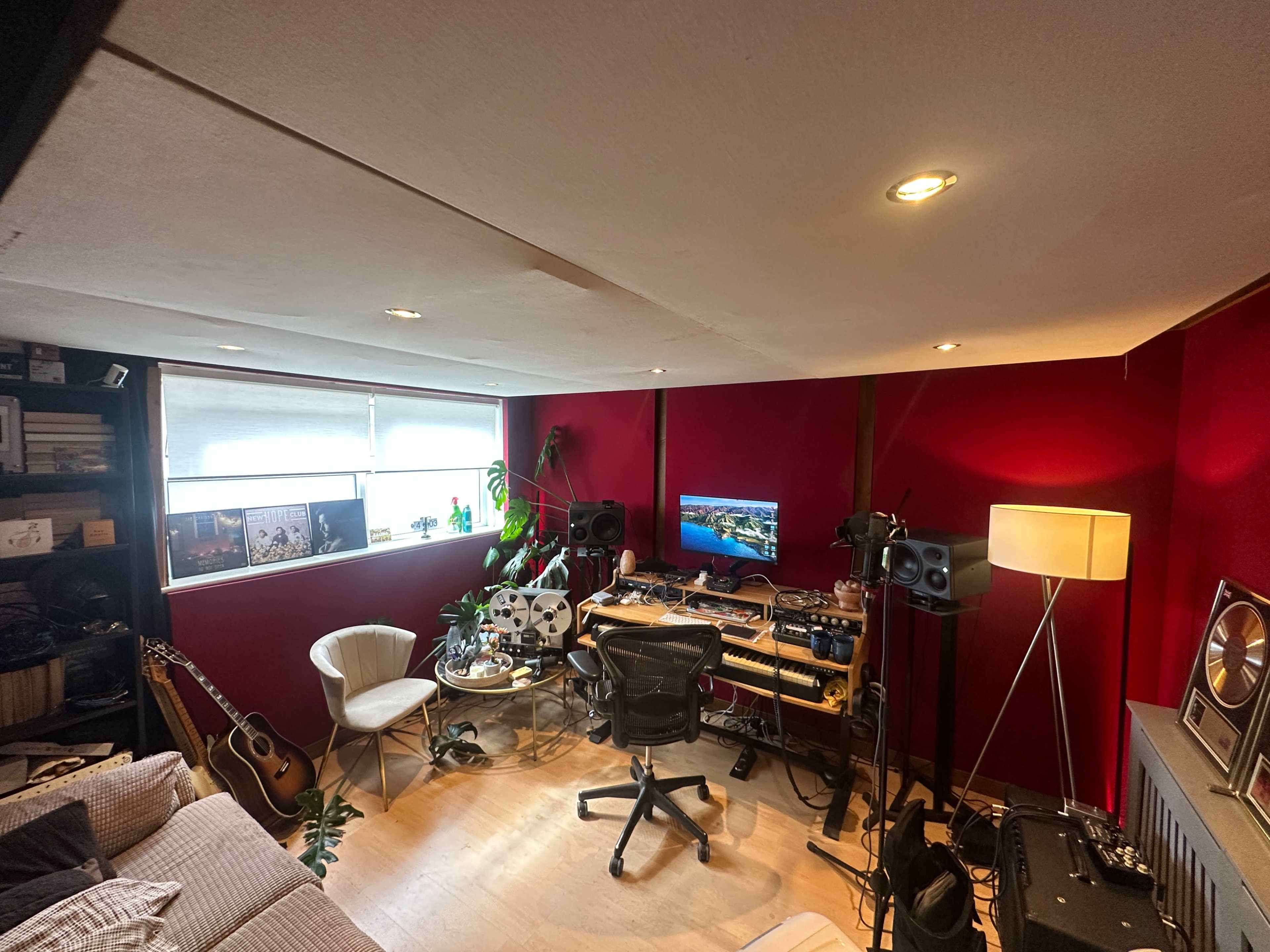 The image shows a home studio with a desk equipped with audio equipment, a computer monitor, and plants, surrounded by red walls and a cozy seating area.