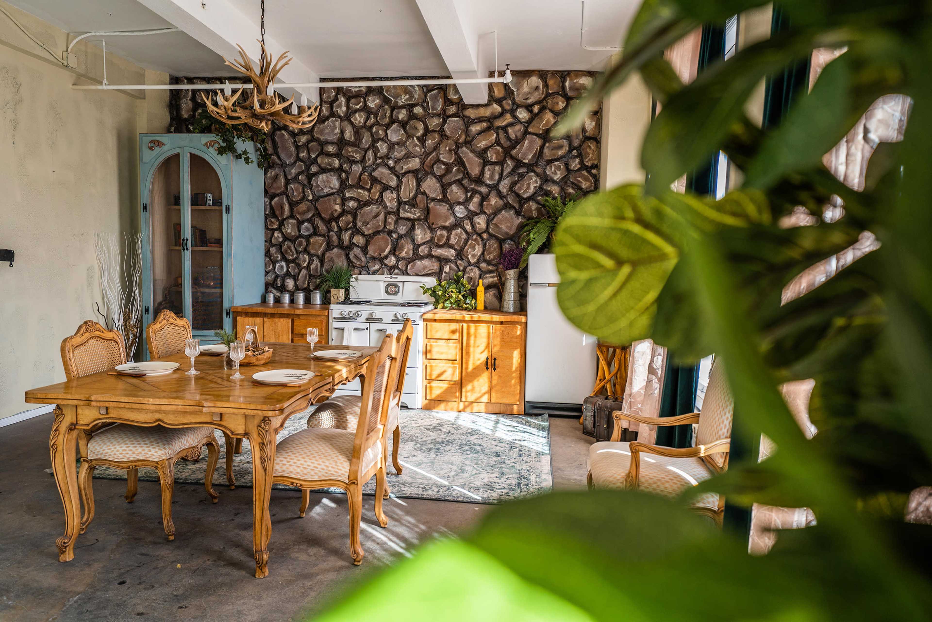 A rustic dining area features a wooden table set for a meal, with a stone wall and vintage appliances in the background.