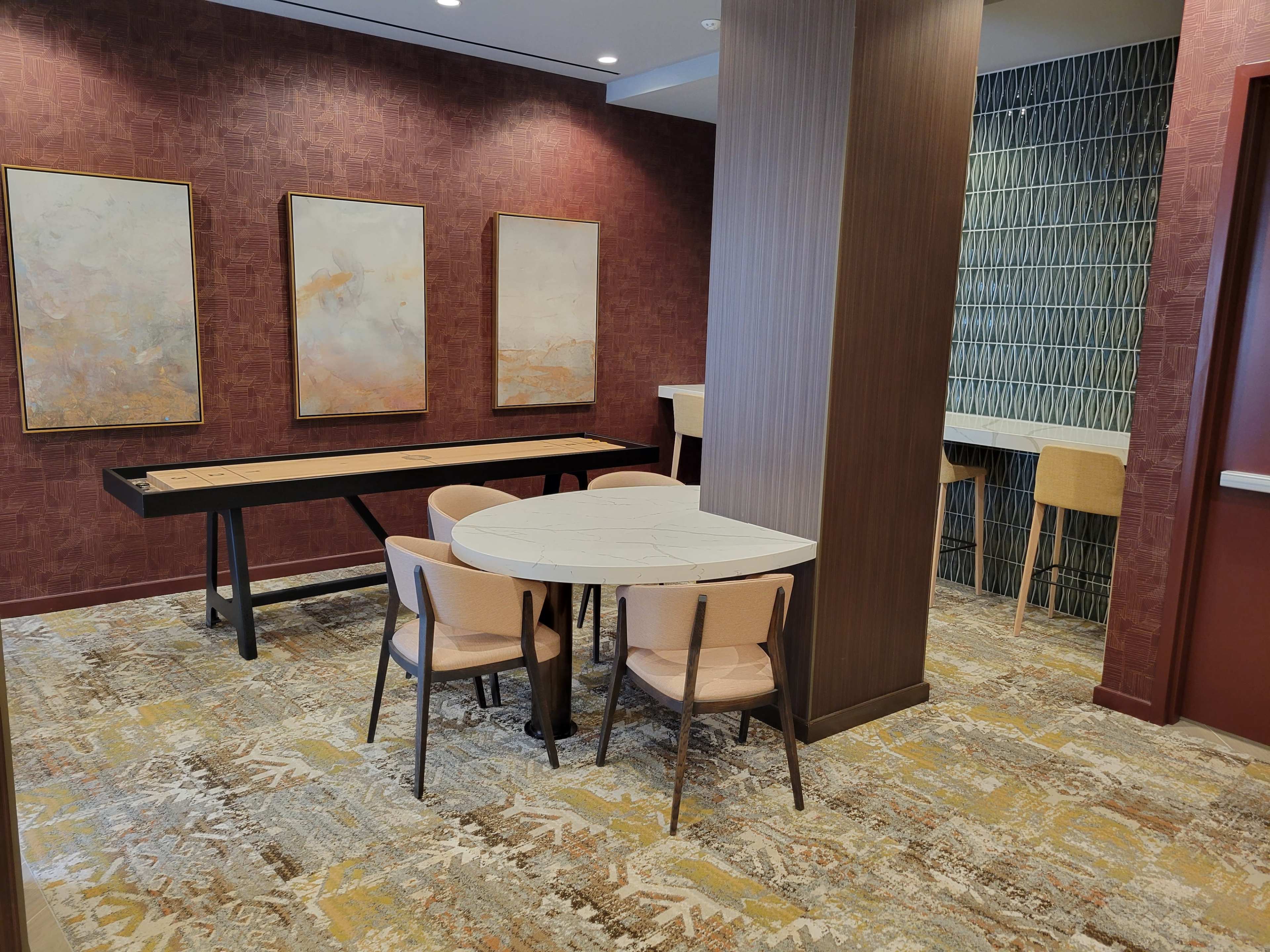 A modern lounge area featuring a small round table surrounded by four chairs, art pieces on the wall, and a long table with high stools in the background.