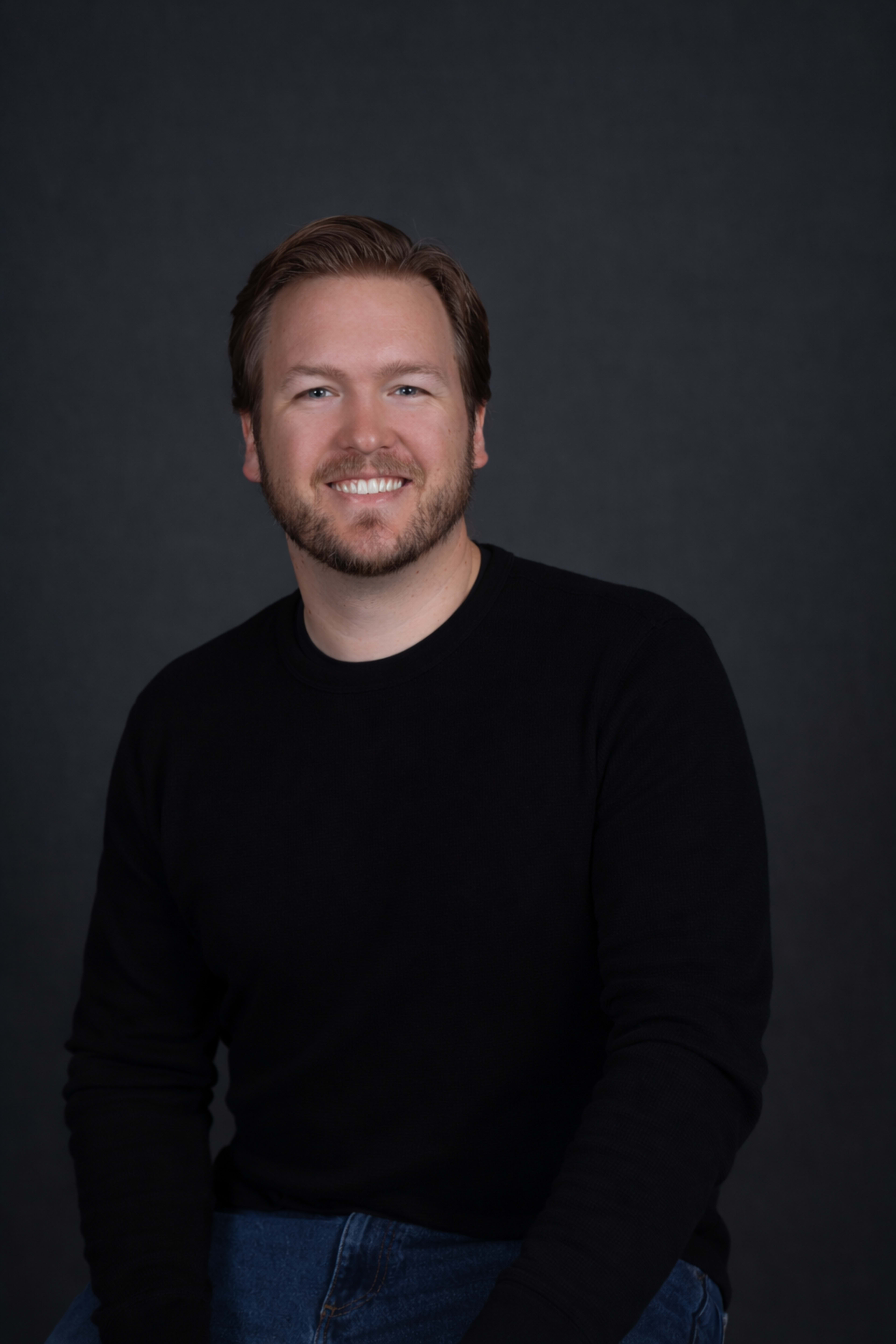 A man with light brown hair and a beard sits against a dark background, wearing a black sweater and blue jeans.