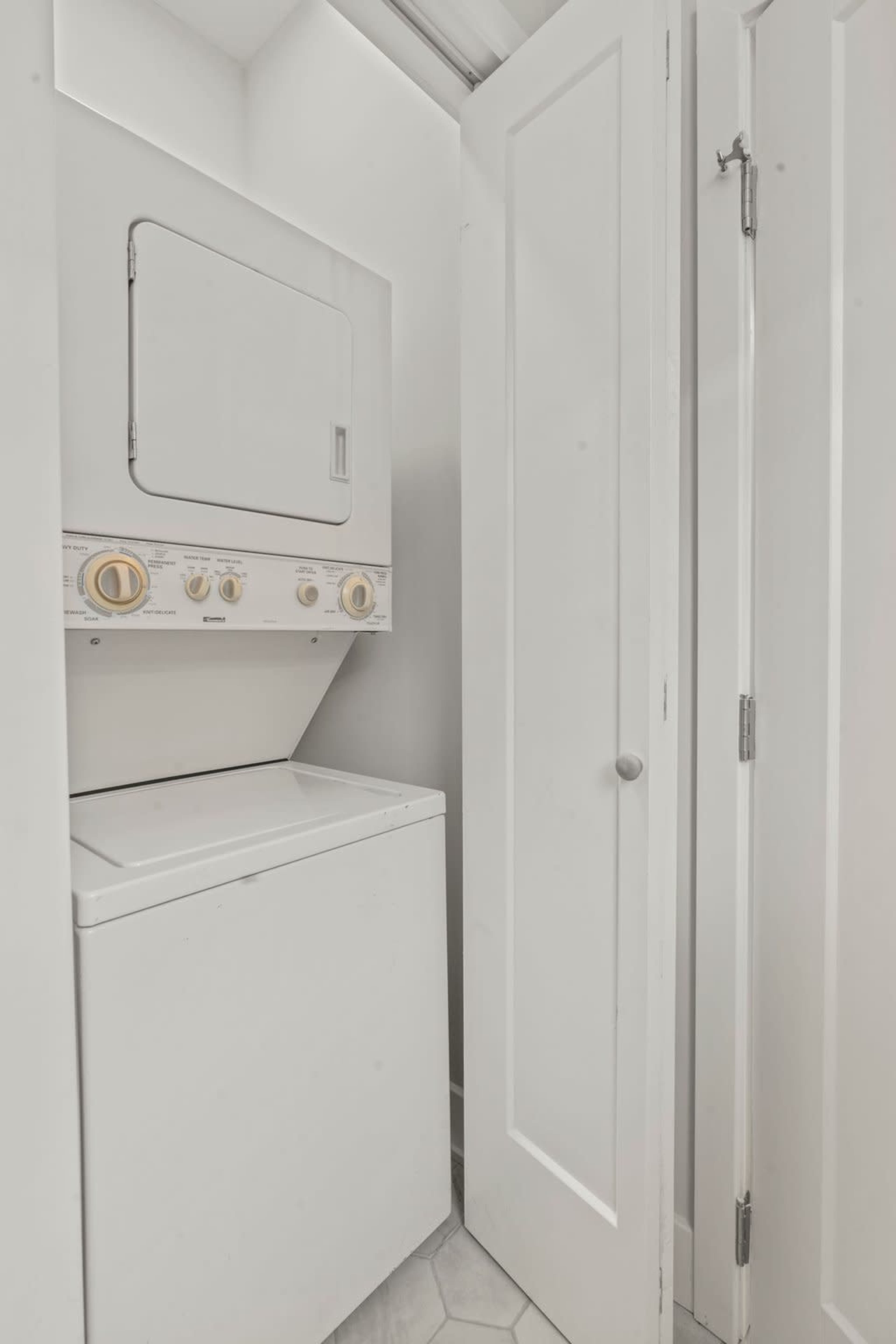 The image shows a compact laundry area with a stacked washer and dryer next to a closed white door.