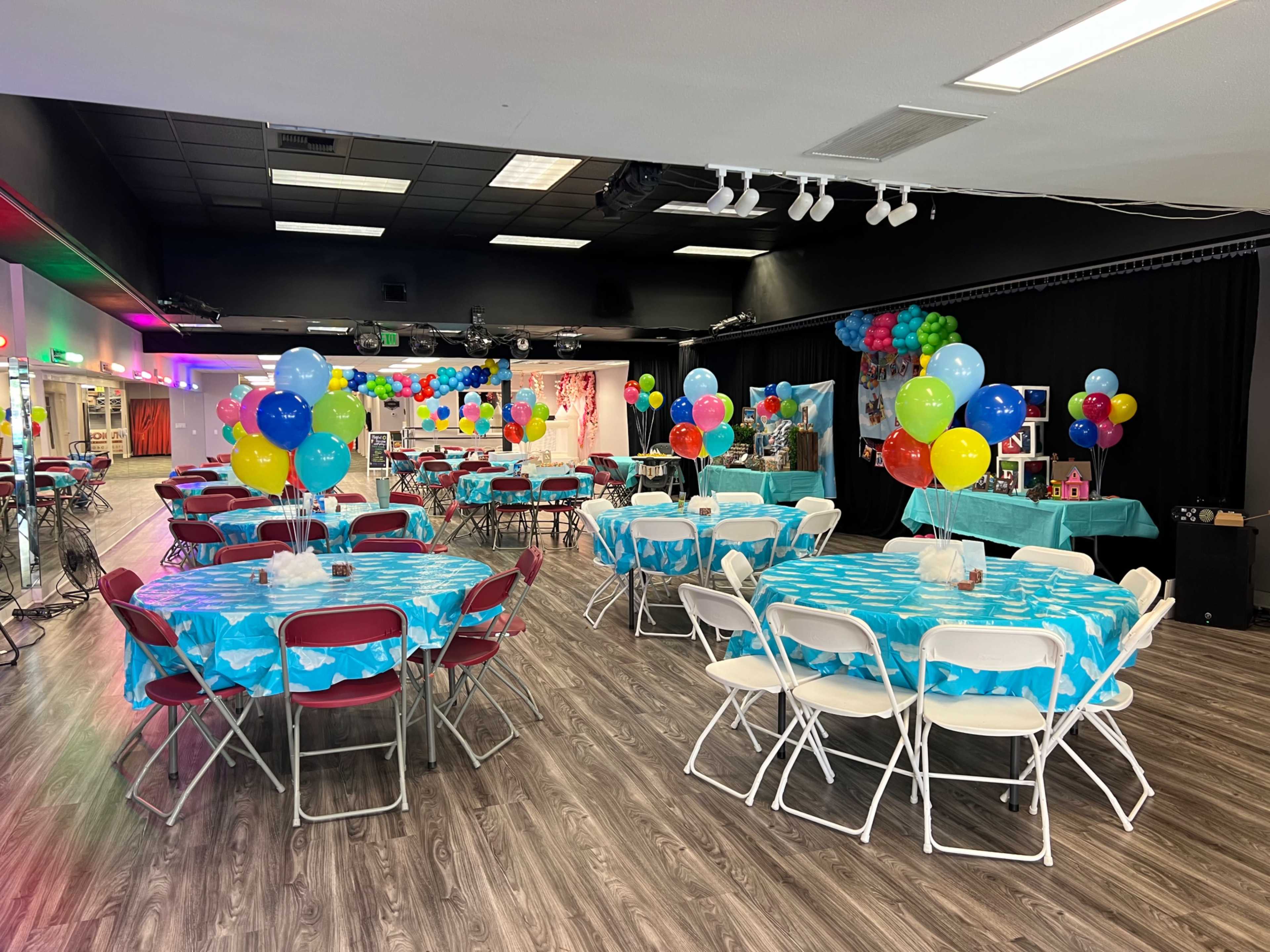 A brightly decorated event space features several tables covered in turquoise tablecloths, surrounded by colorful balloons and festive decorations.