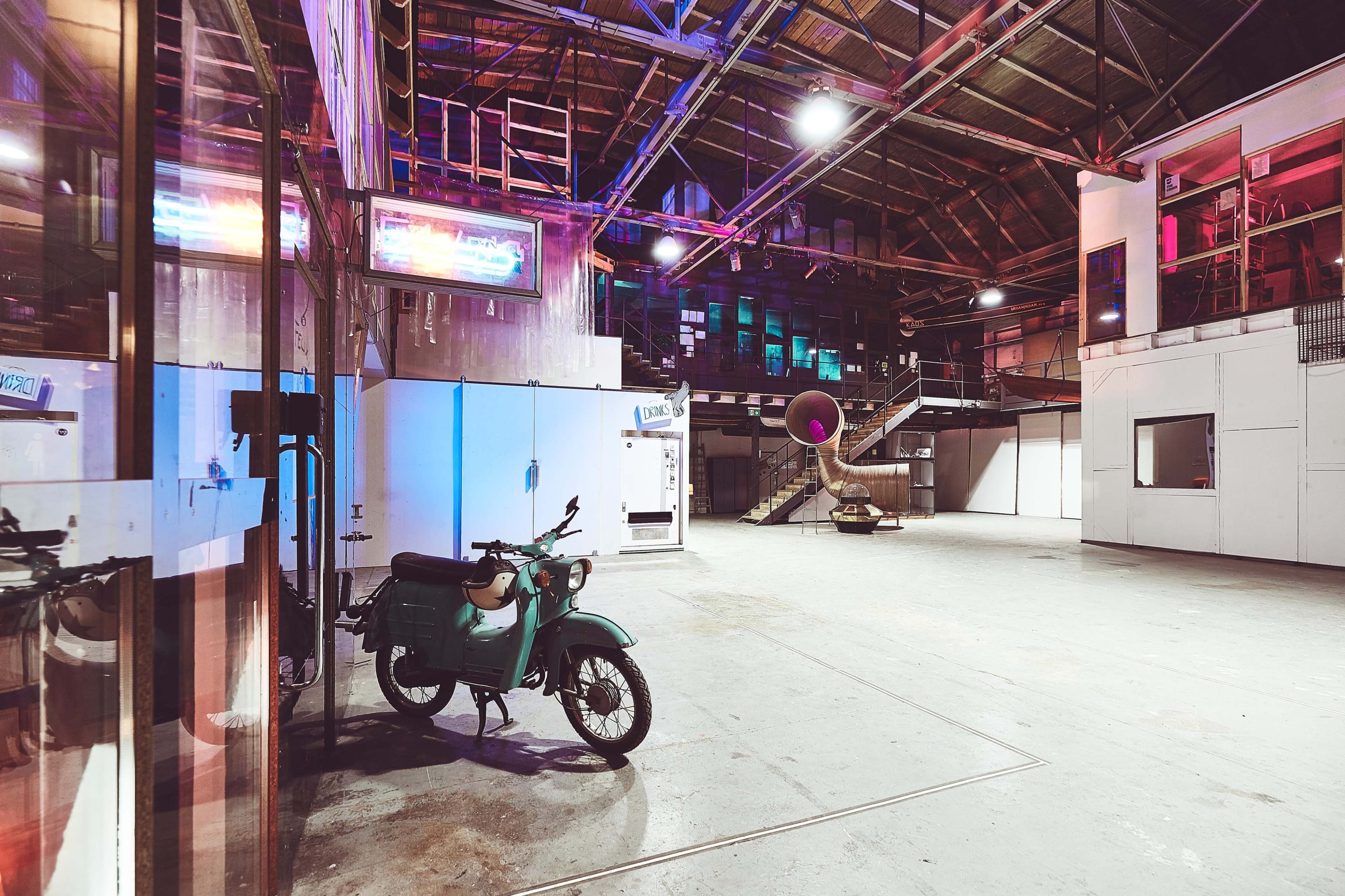 An empty industrial space with a green motorcycle parked in front and a staircase leading to an upper level illuminated by colorful lights.