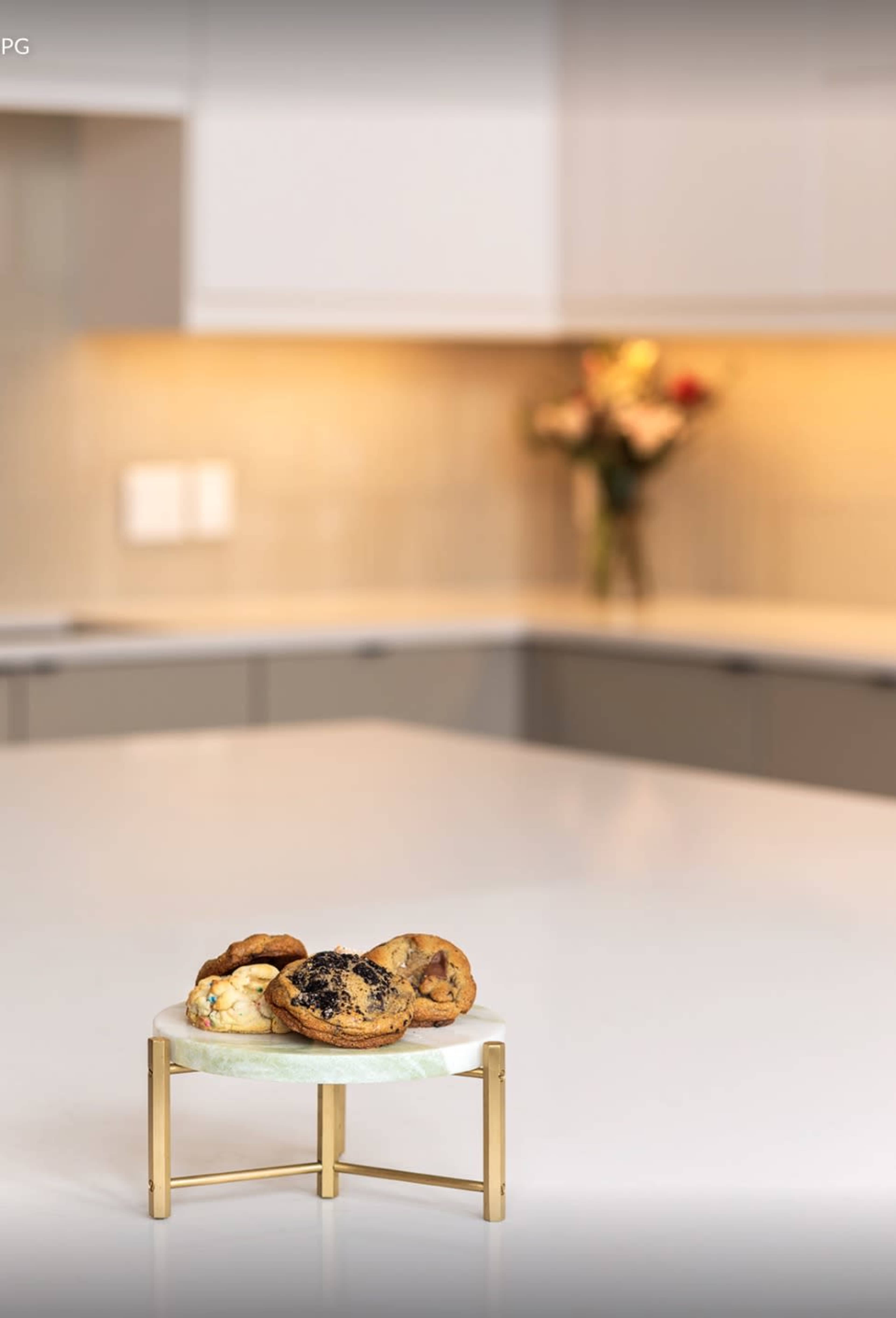 A white marble cake stand holds a variety of baked goods on a kitchen countertop with blurred flowers in the background.