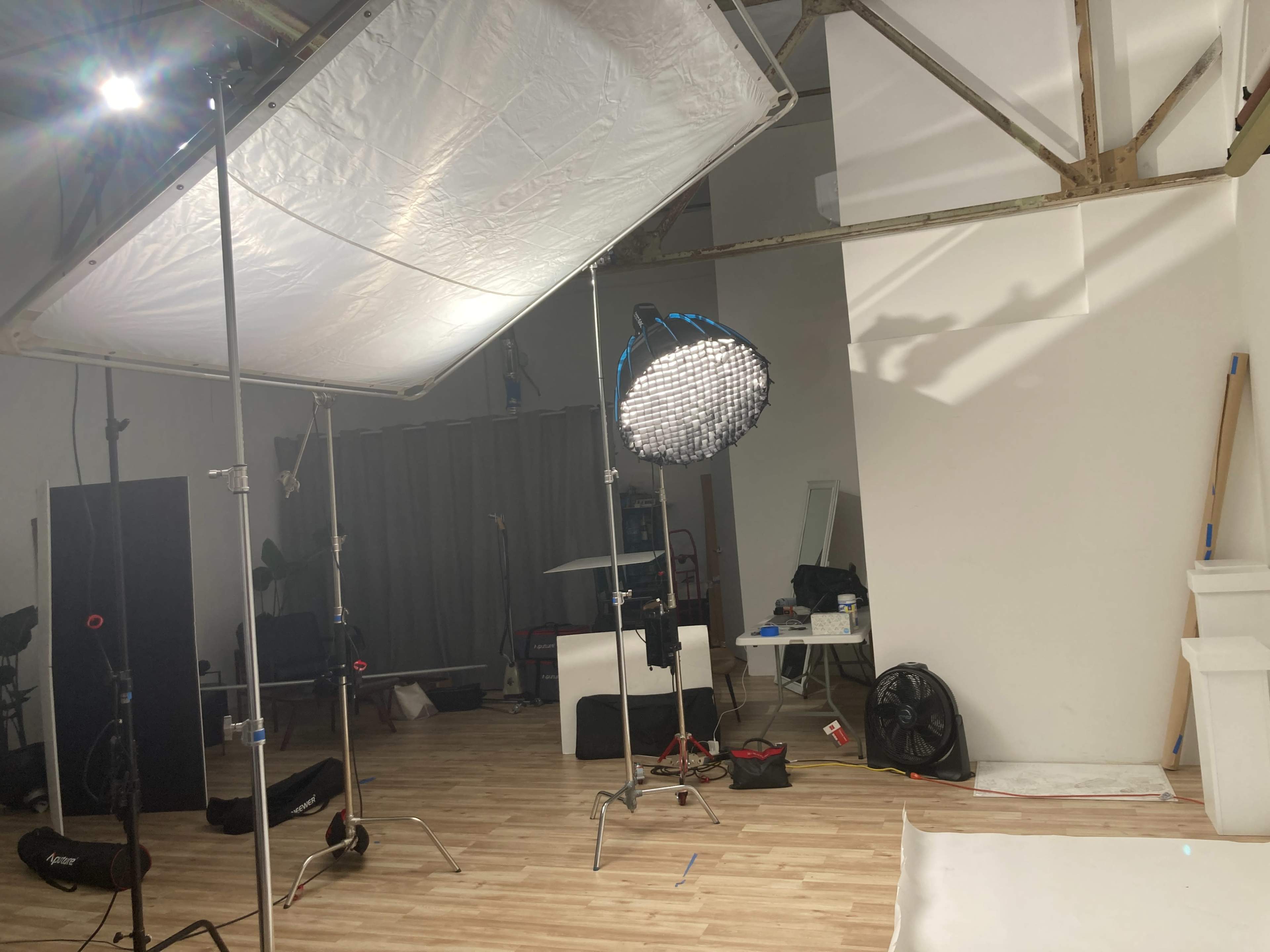 The image shows a photography studio set up with lighting equipment, a large softbox, and a circular light fixture in a room with wooden flooring and various studio props scattered around.