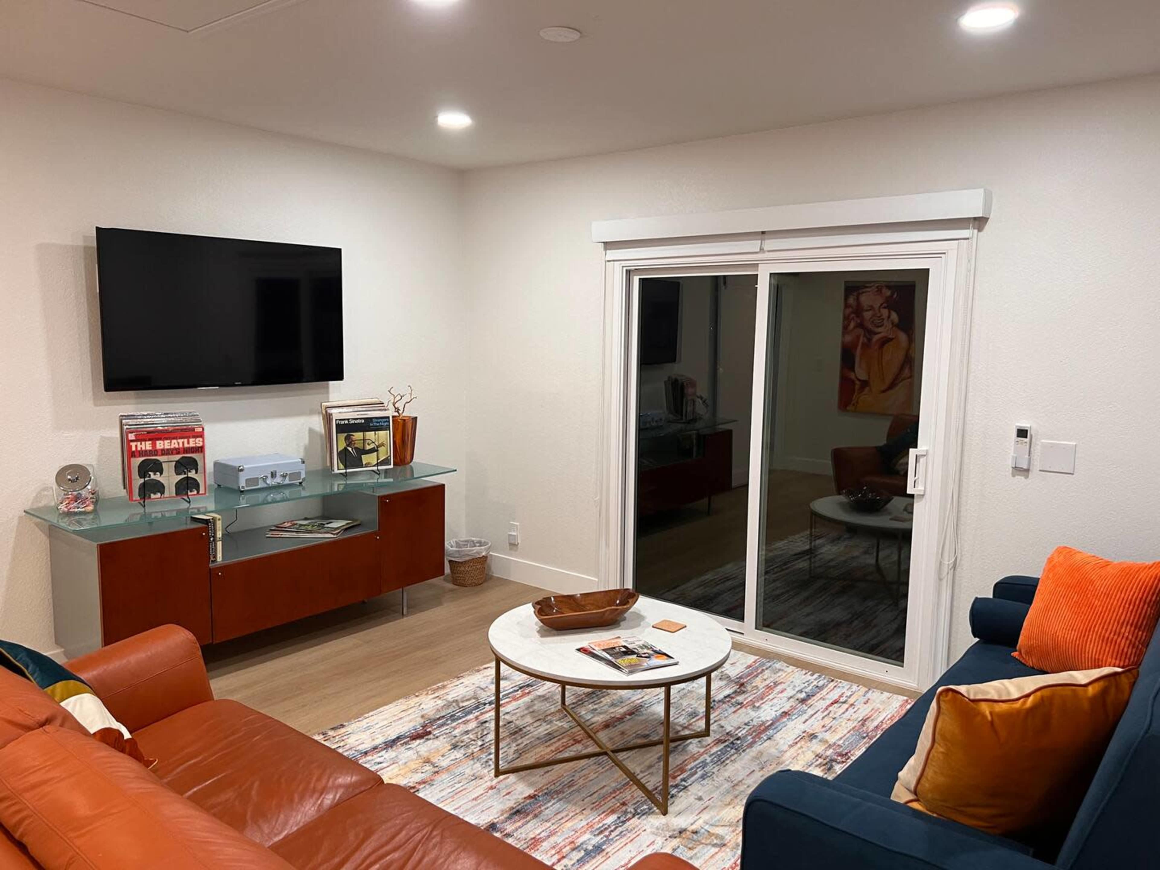 The room features a modern living area with a television mounted on the wall, a glass coffee table, and a sliding door leading outside, surrounded by a stylish couch and various decor items.