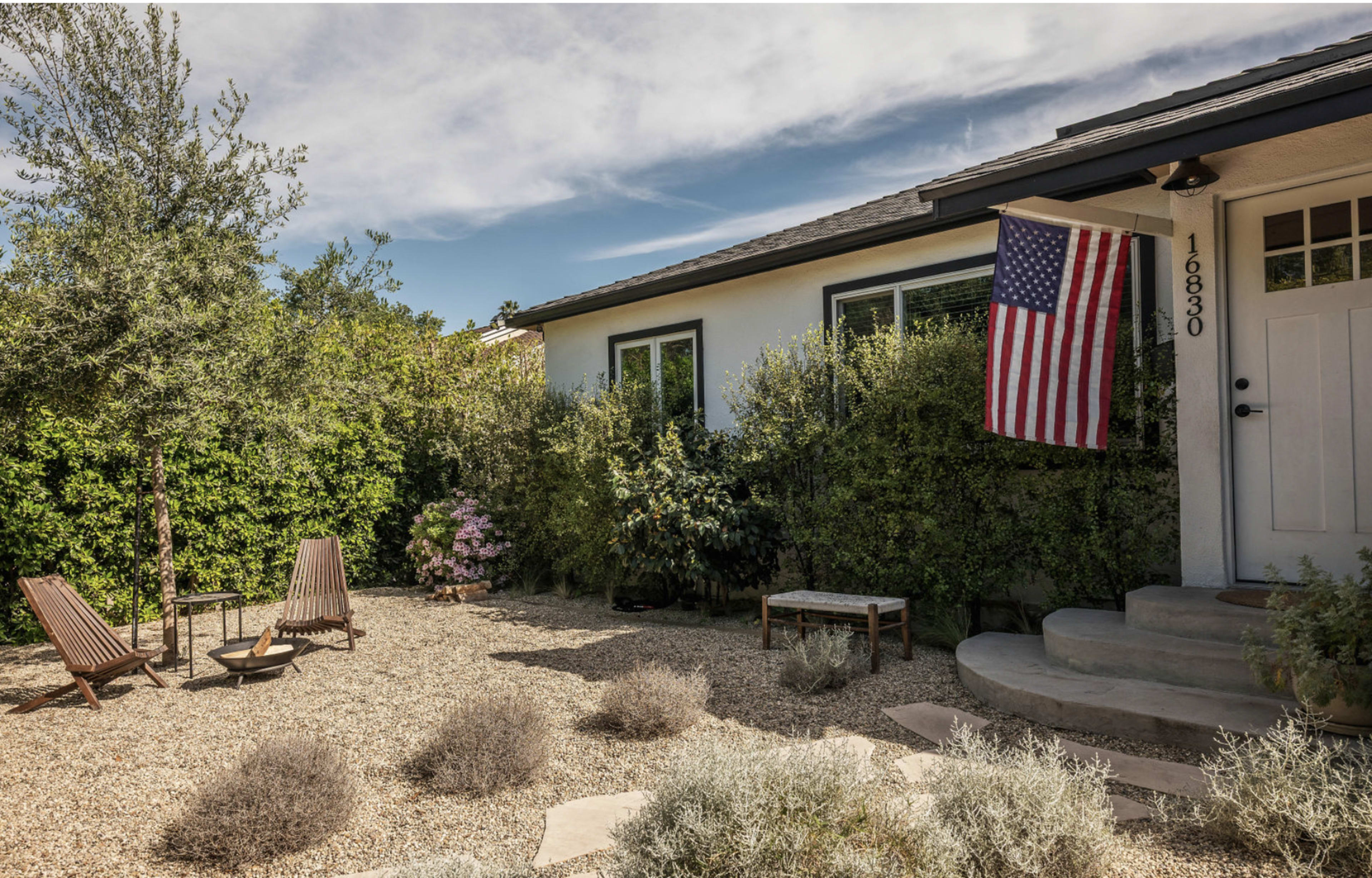 A small, gravel-covered yard features two lounge chairs and a table, with a front door and an American flag hanging beside it.