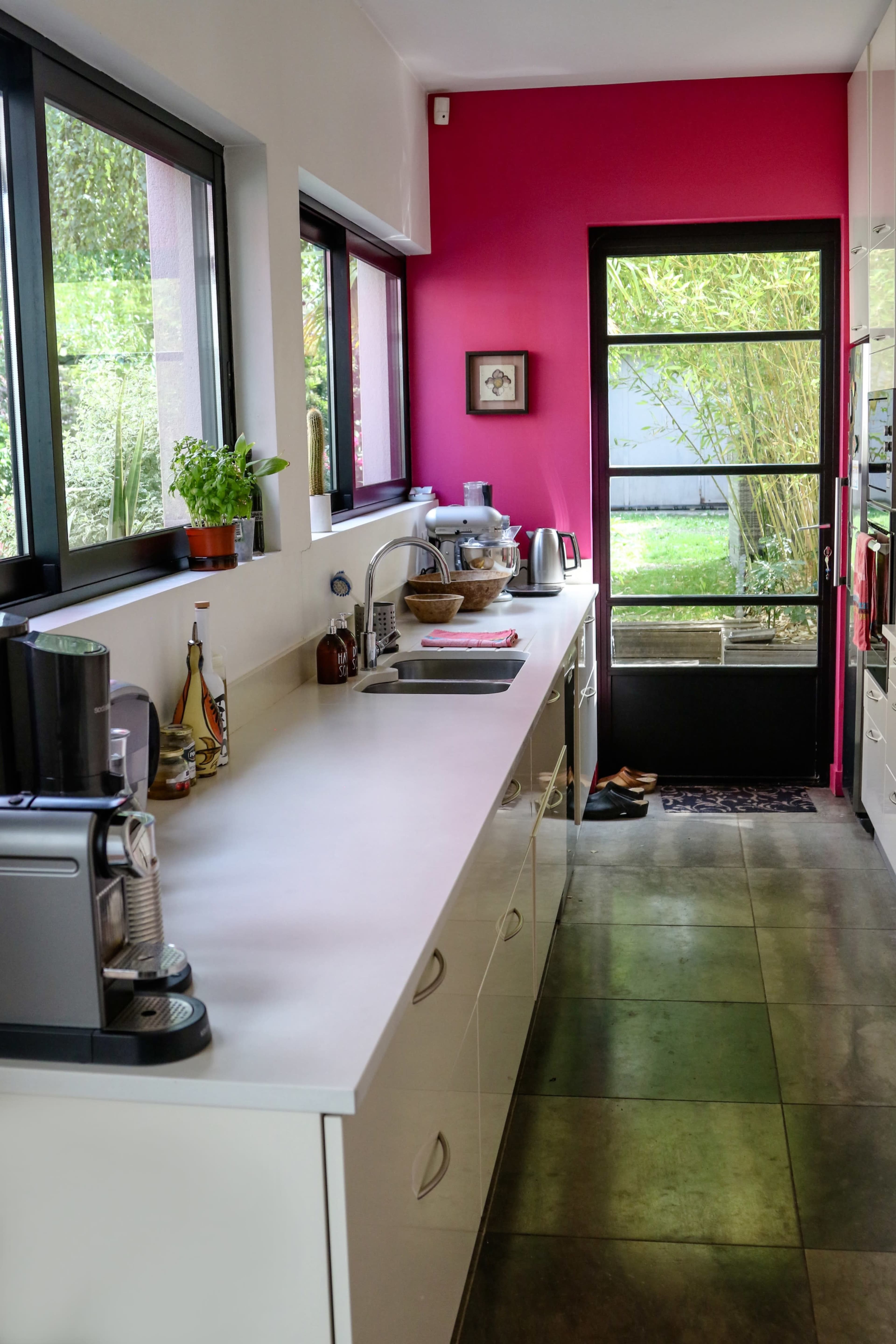 A narrow kitchen with a sleek countertop, modern appliances, and a vibrant pink accent wall, leading to a glass door.