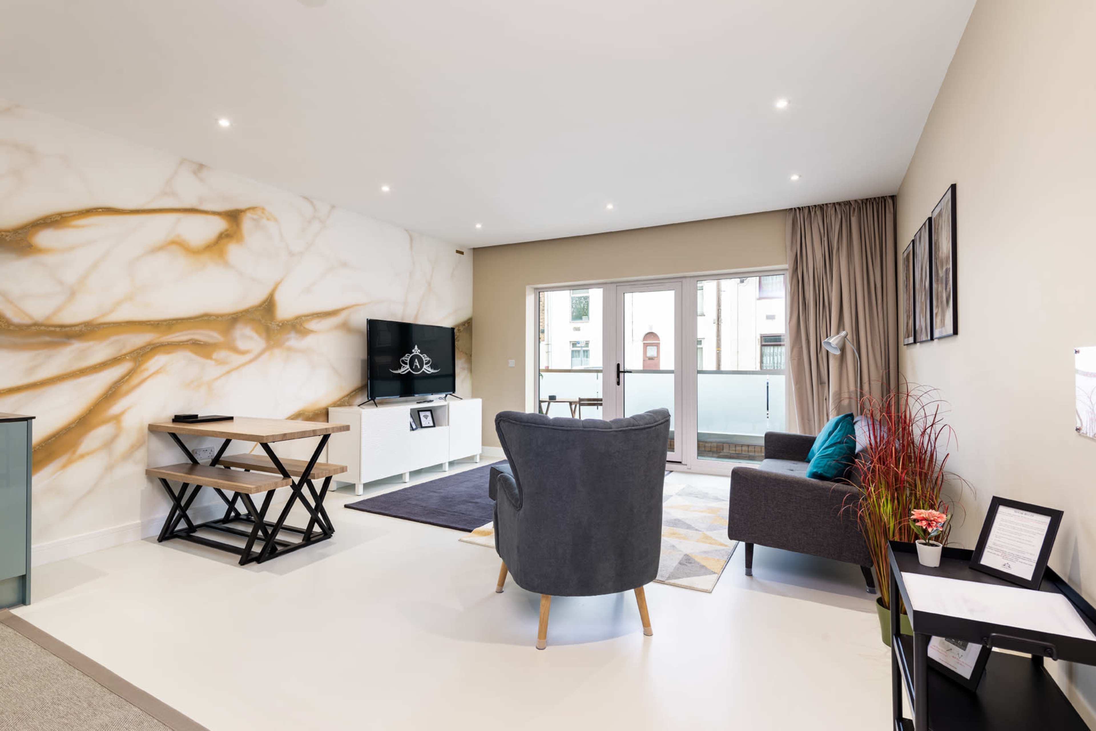 A modern living room with a distinct marble wall, a television mounted on a white cabinet, a gray armchair, a sofa, and a small dining table.