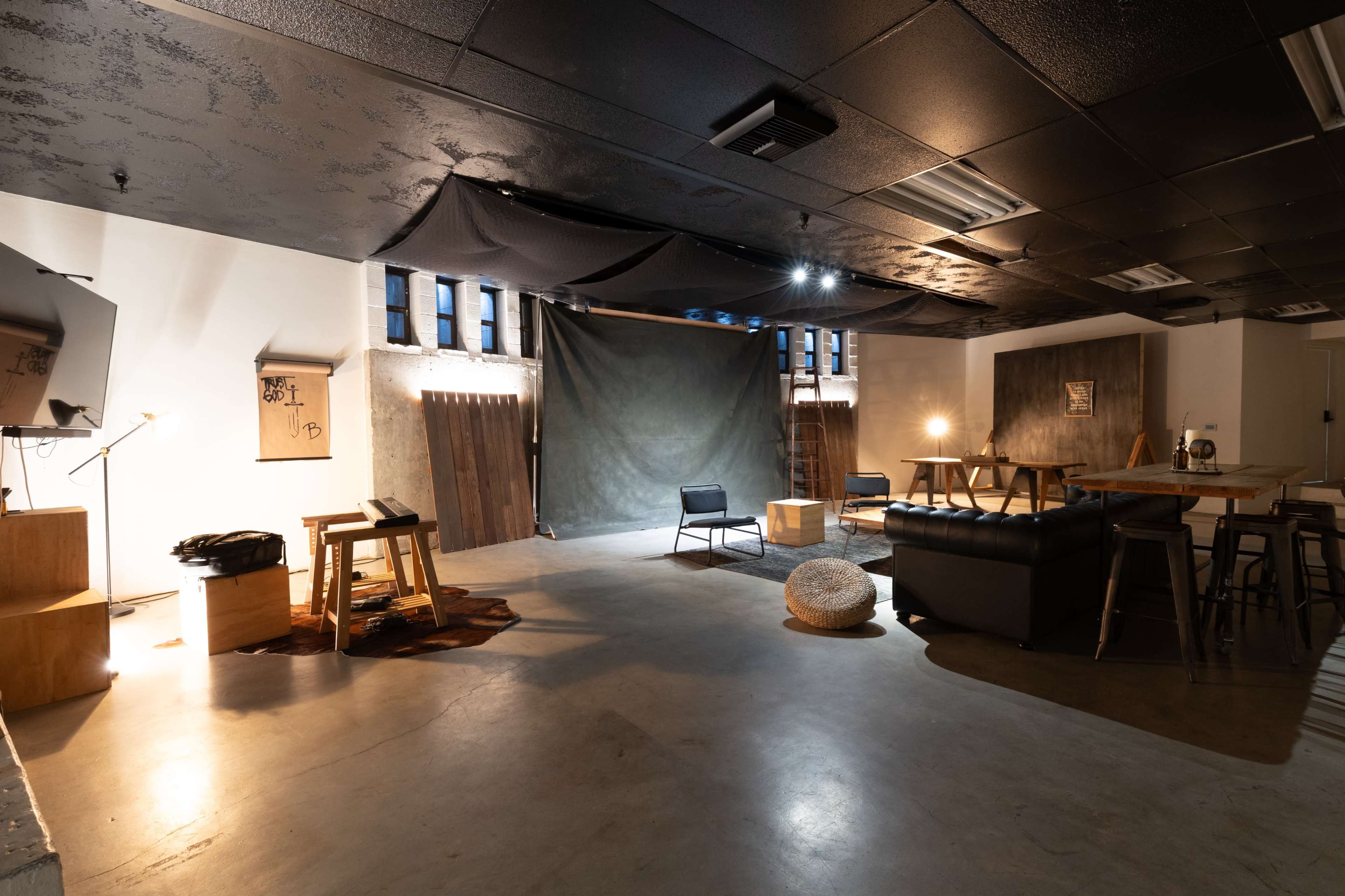 The image shows a spacious, dimly-lit room with various seating areas, a wooden table, a couch, and a backdrop setup, featuring industrial-style decor.