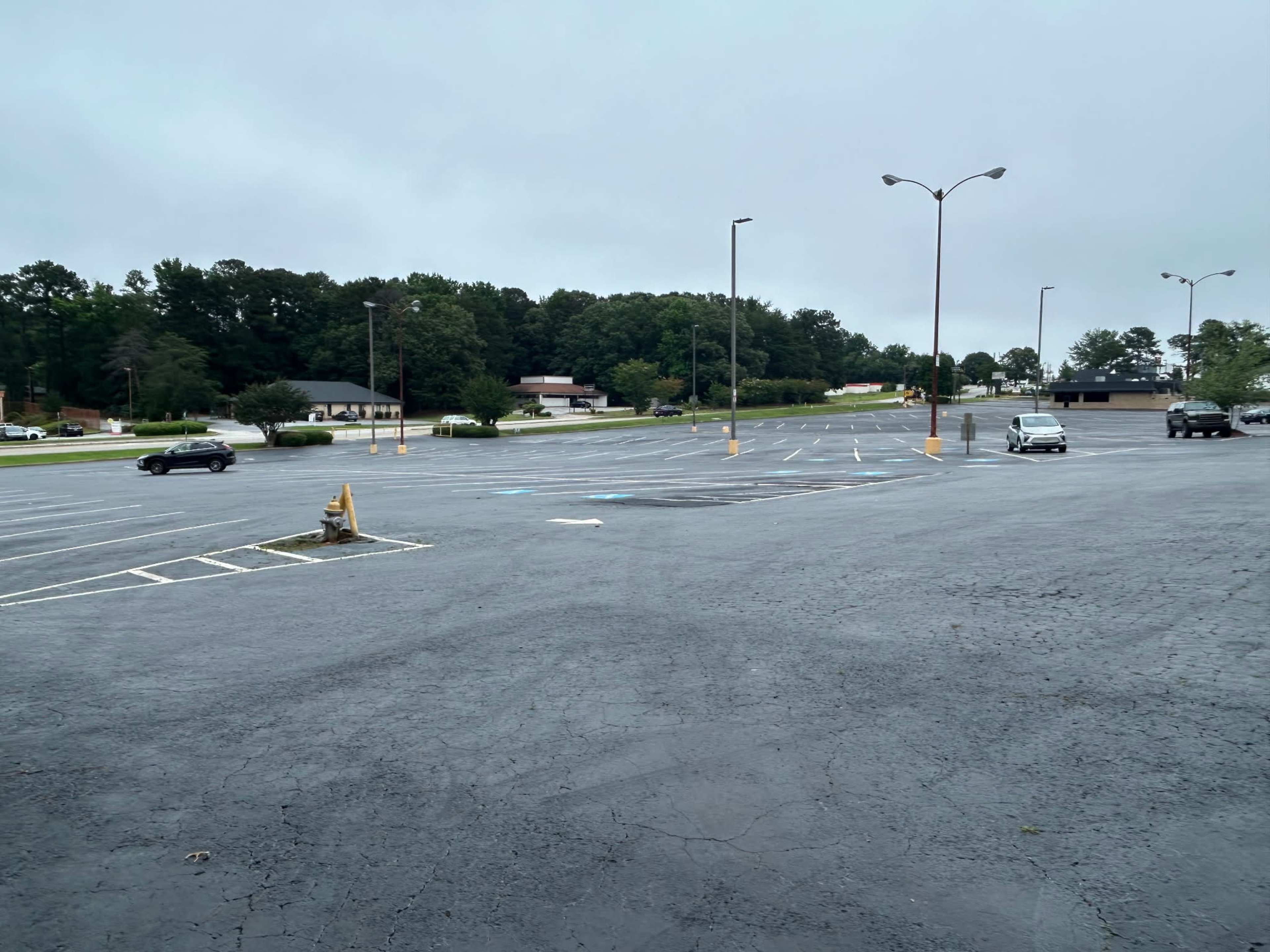 A nearly empty parking lot shows several parked cars and a few trees in the background under a cloudy sky.