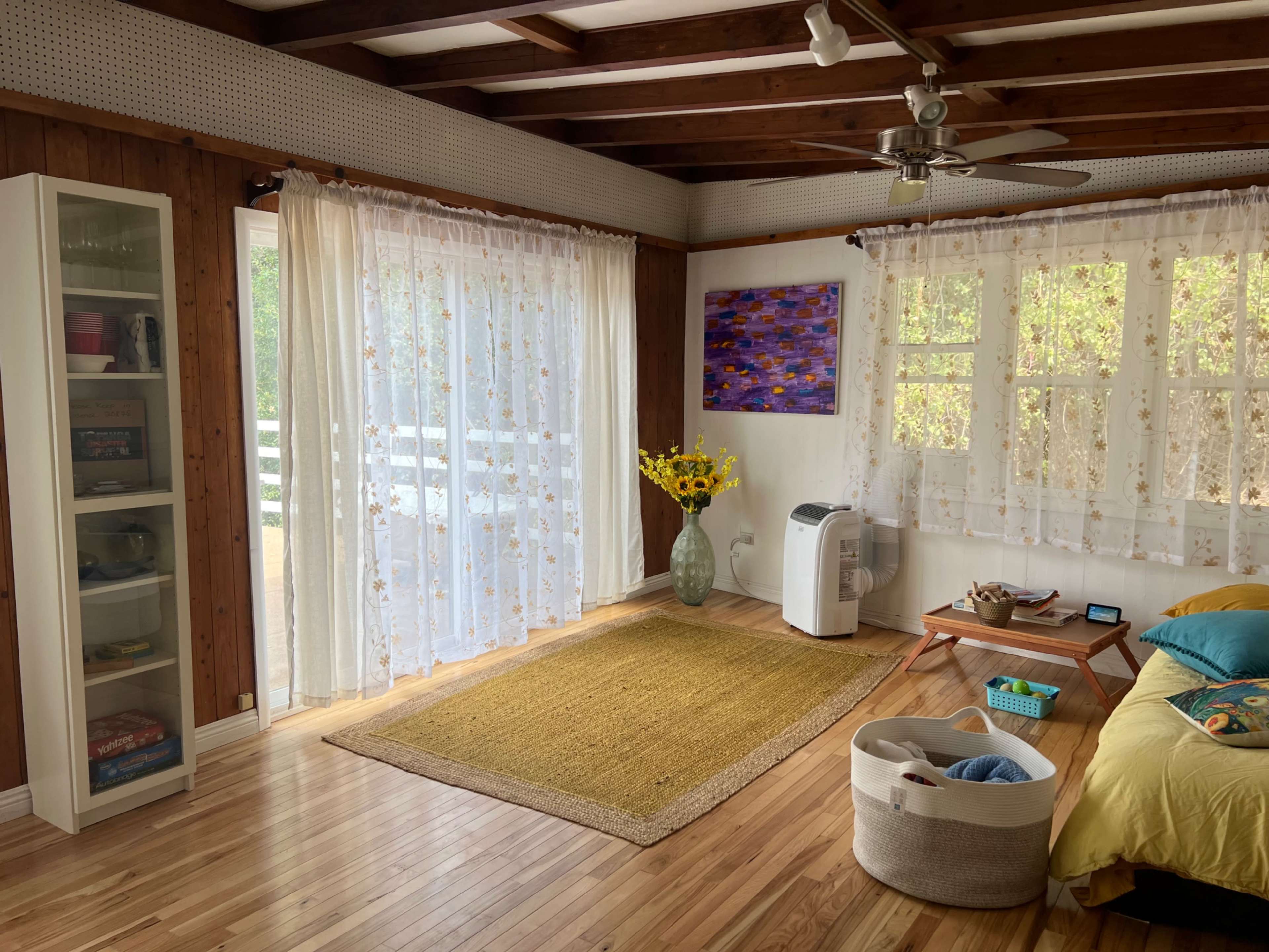 A bright living room features wooden walls, a light-colored rug, a couch with colorful cushions, and large windows covered with sheer curtains.