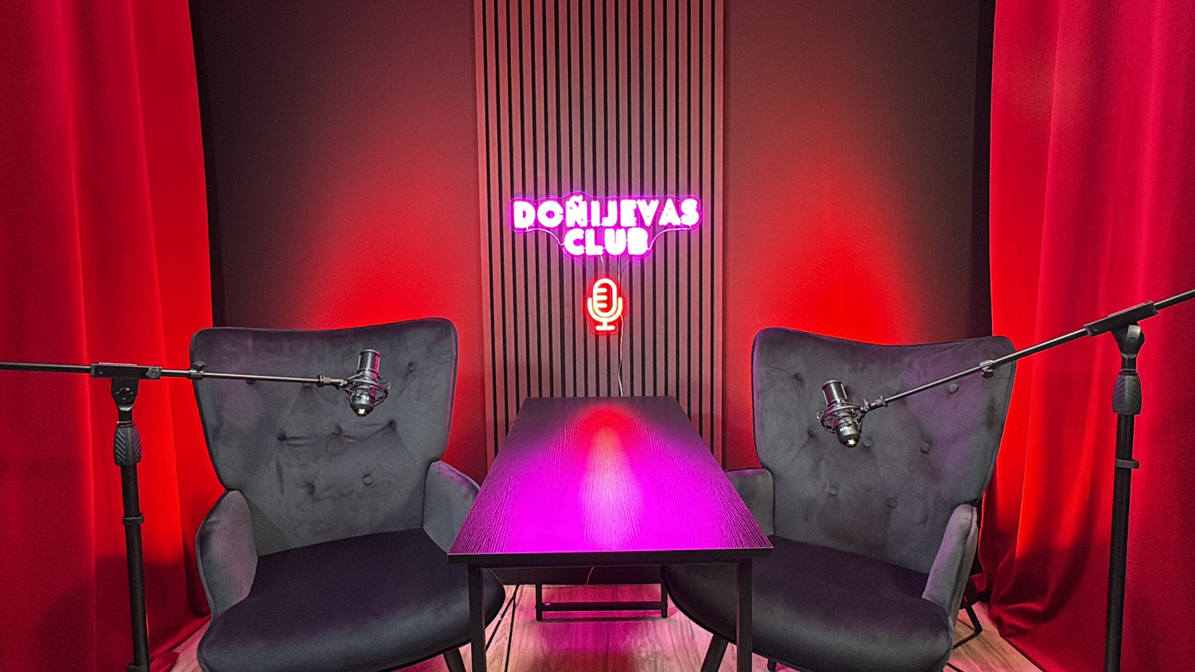 The image shows a podcast or interview setup featuring two chairs with microphone stands and a table, illuminated by red lighting and a neon sign that reads "DOJILJEVAS CLUB."