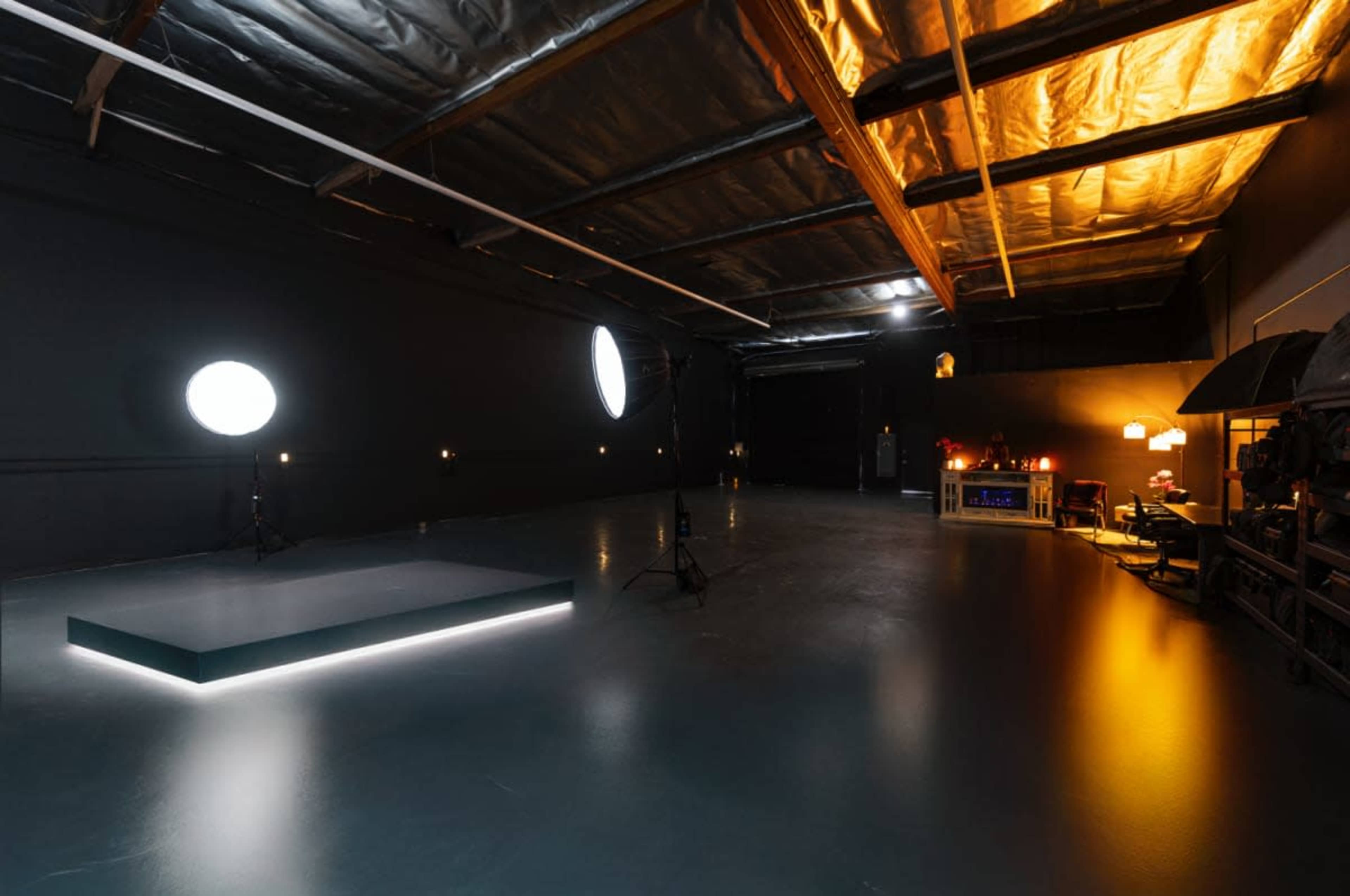 The image shows a spacious studio with a dark, polished floor, a small illuminated platform in the center, and various lighting equipment and furniture arranged along the walls.
