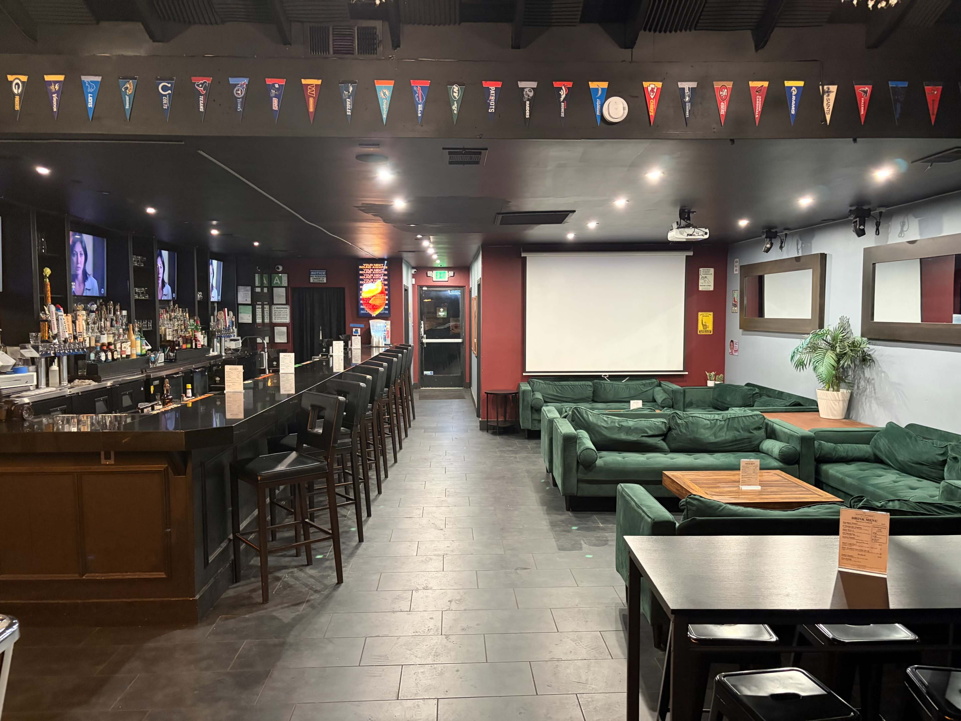The image shows a dimly lit bar area with a long counter, green sofas arranged along the walls, and a large screen at the front of the room.