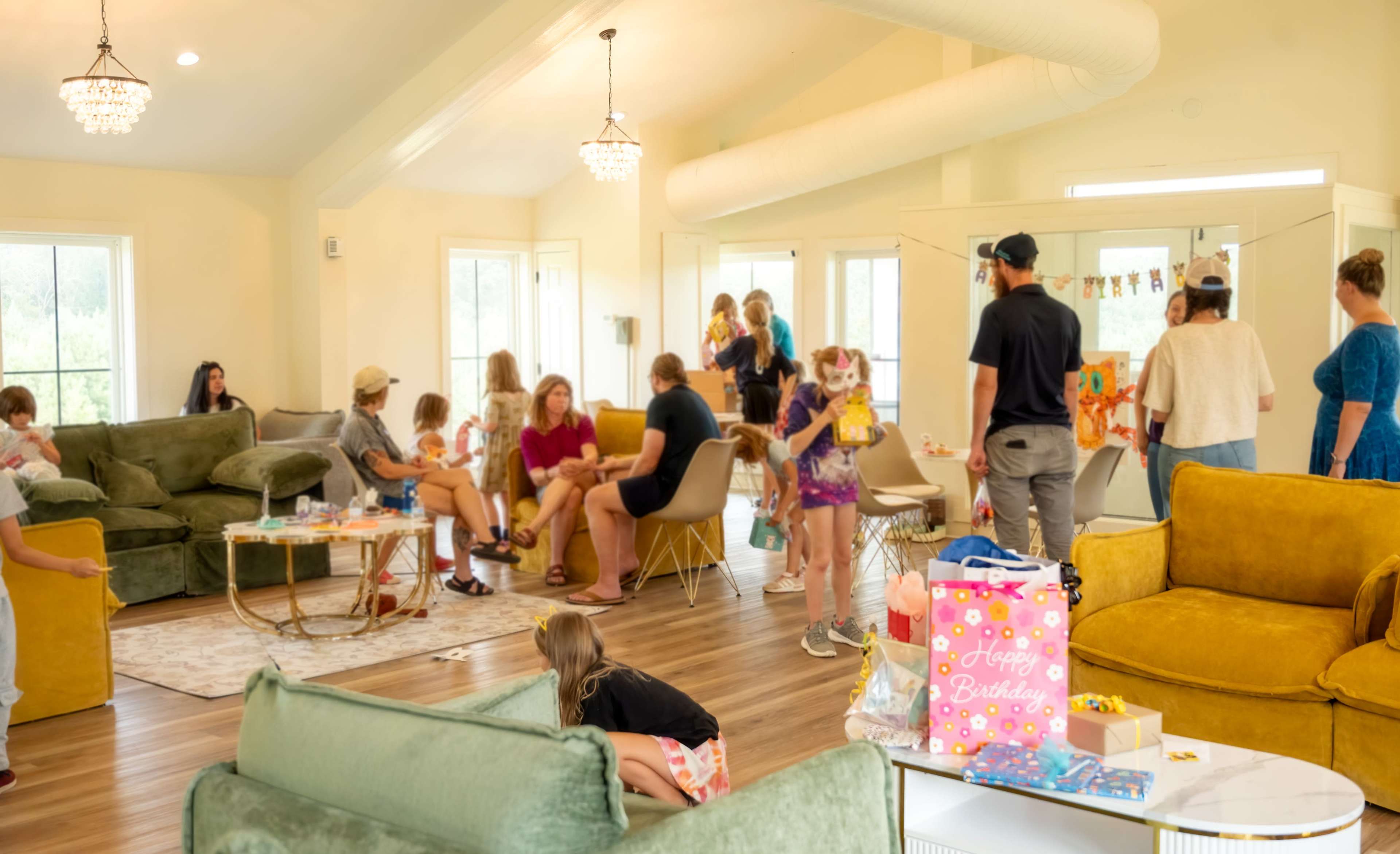 A colorful birthday party is taking place in a bright, spacious room filled with children and adults, with decorations and gifts scattered throughout.