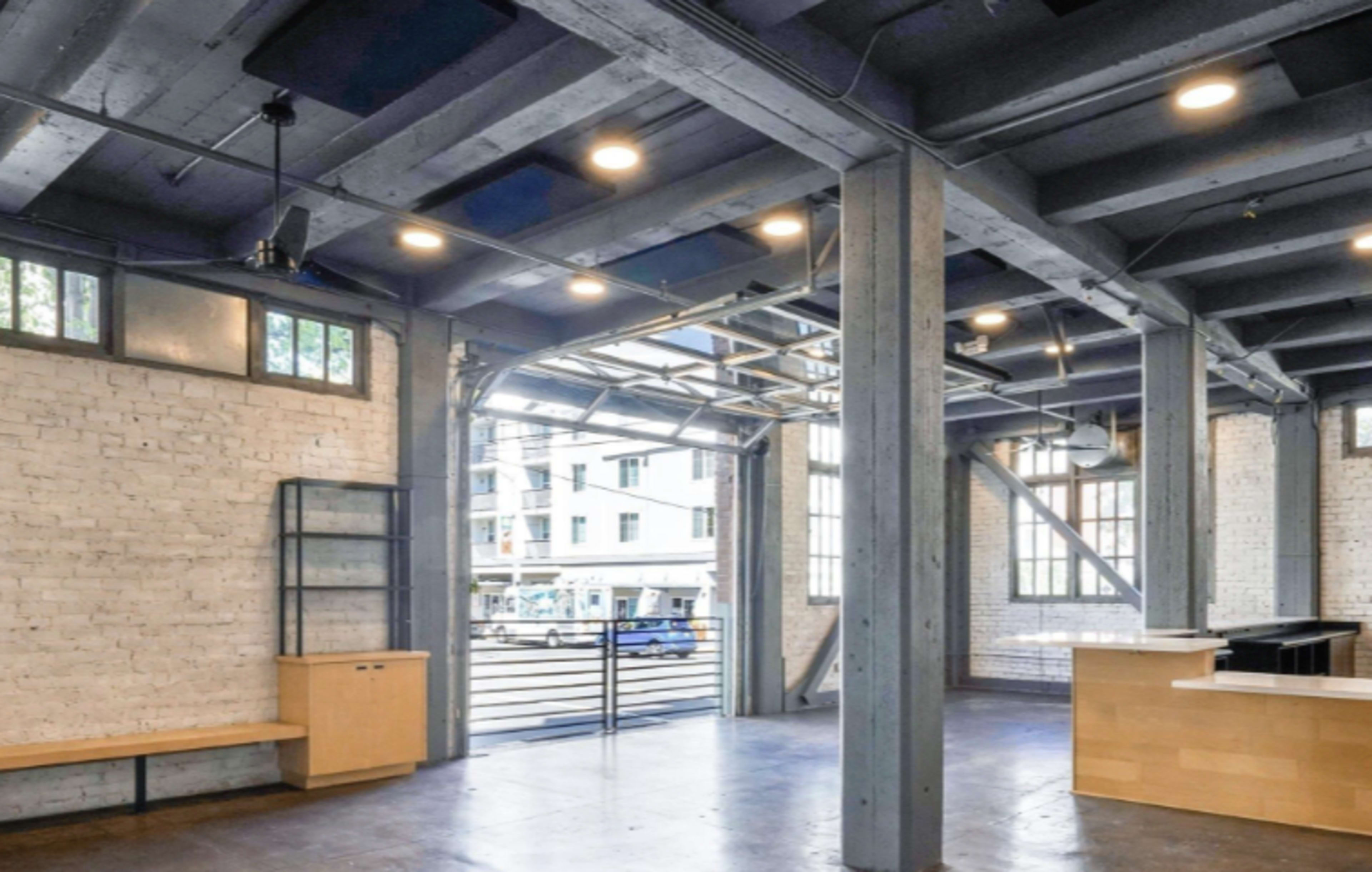 The image shows an industrial-style interior space with exposed beams, brick walls, and large windows, featuring a kitchen area and an open layout.