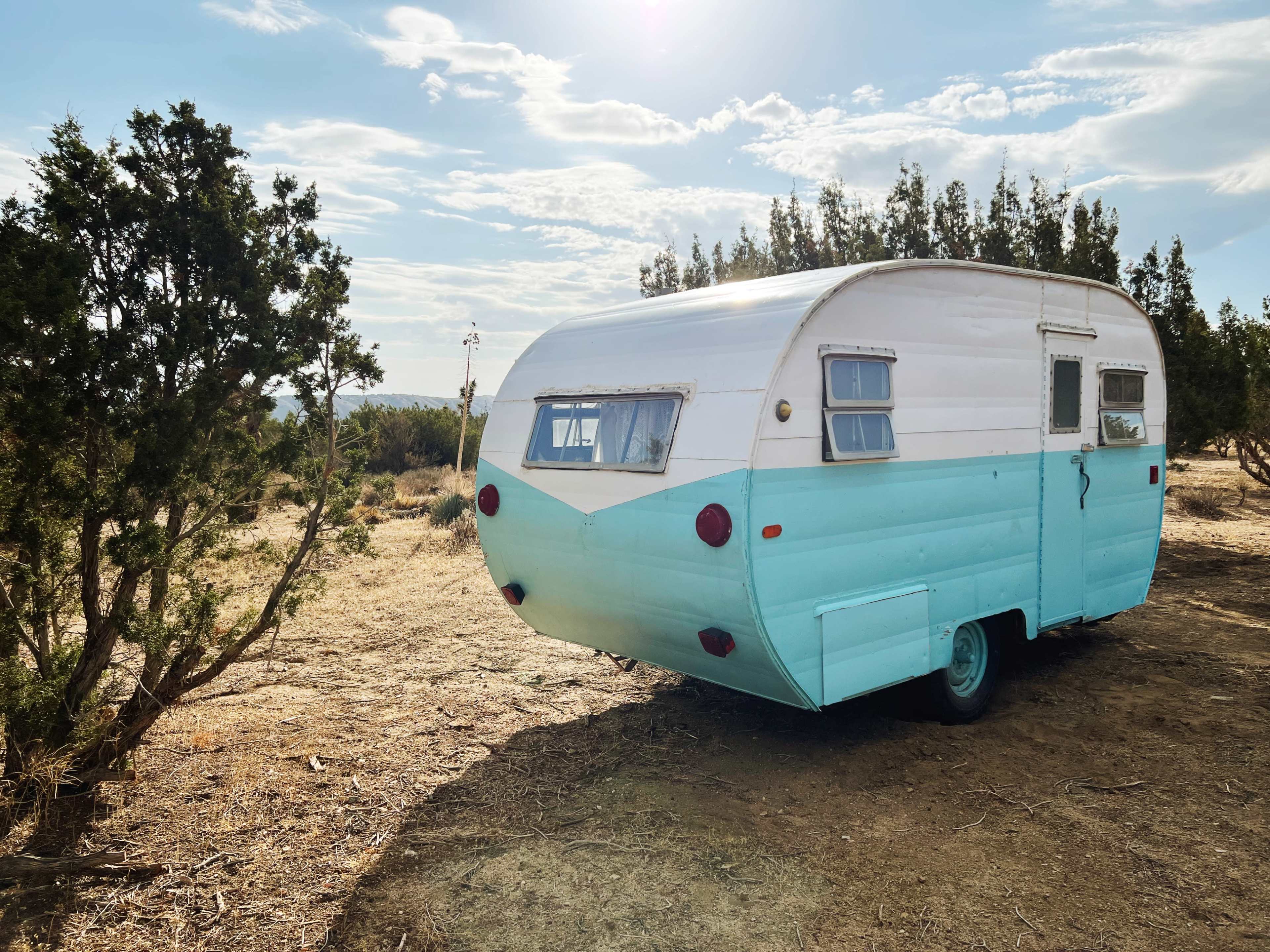 A turquoise and white vintage camper is parked in a sparsely vegetated area under a cloudy sky.