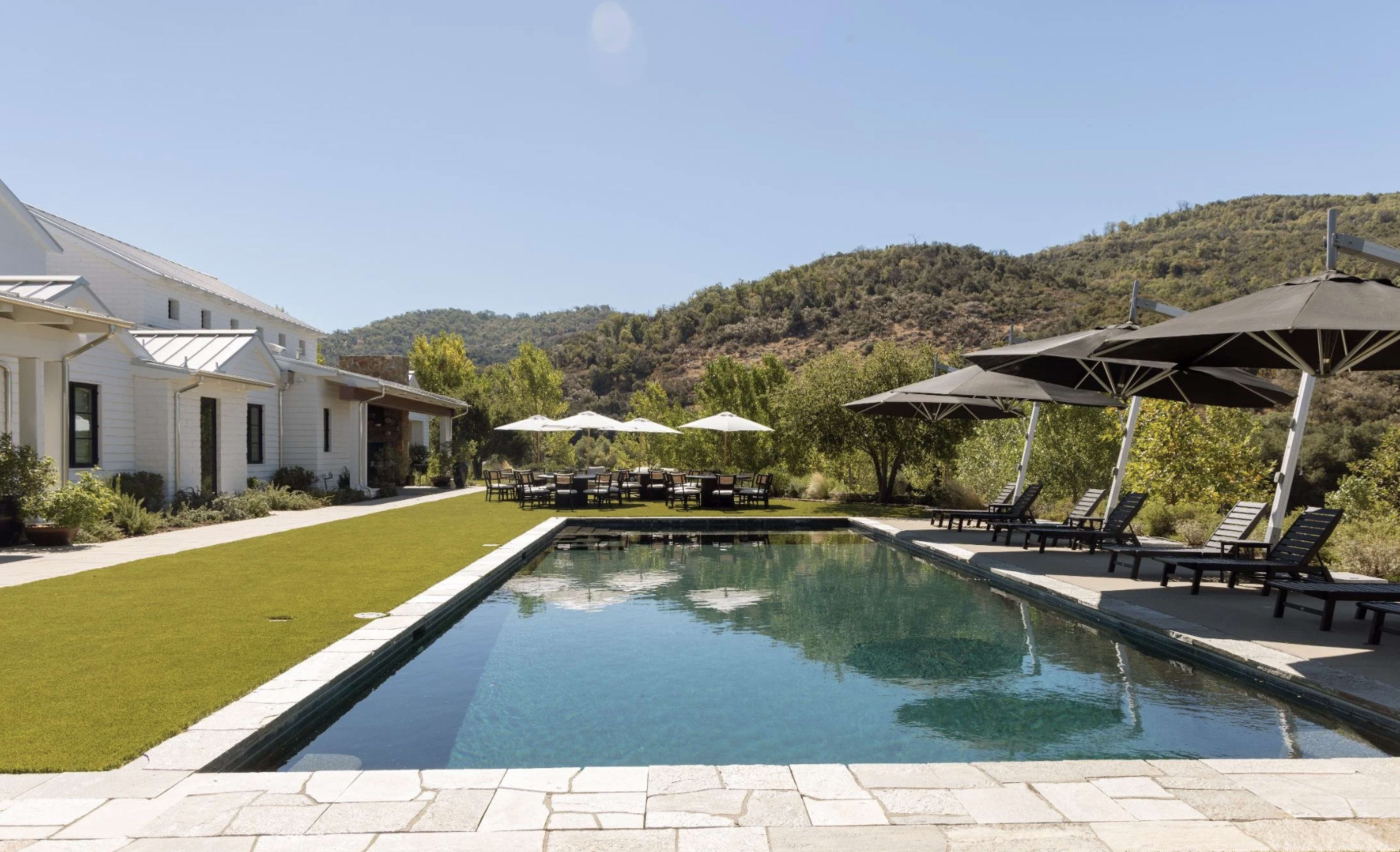 A swimming pool with lounge chairs and umbrellas is situated beside a lawn and a countryside view.