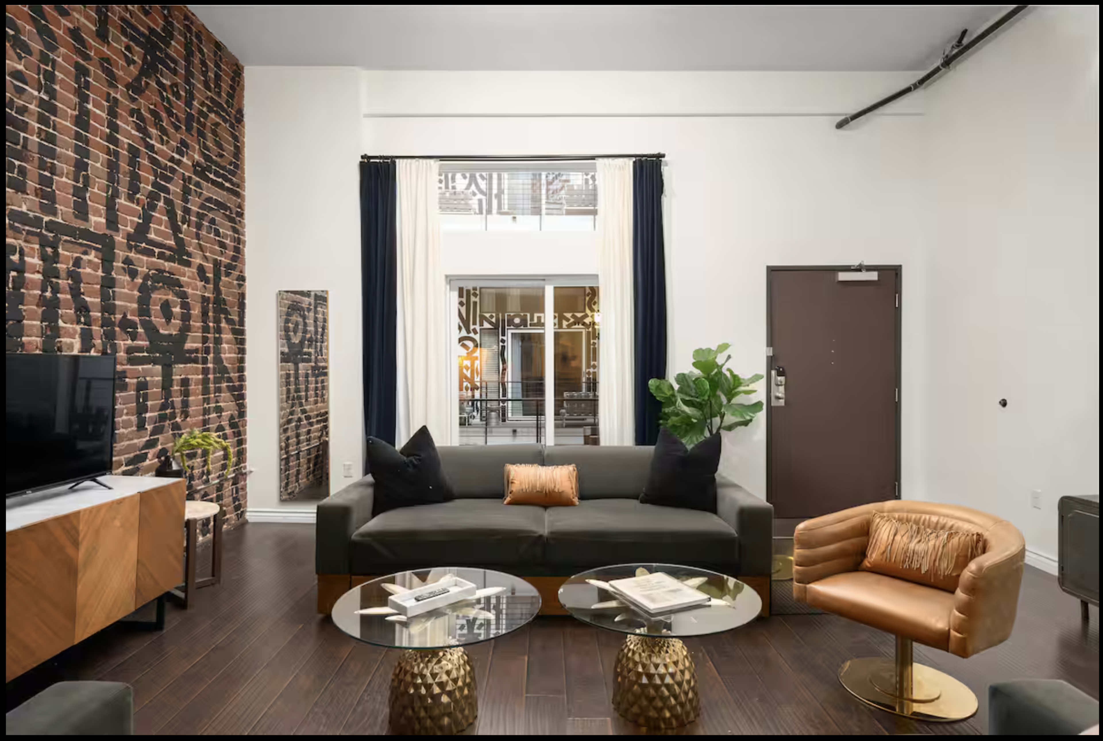 The image shows a modern living room with a brick accent wall, a dark sofa, two round glass coffee tables, and an orange armchair.