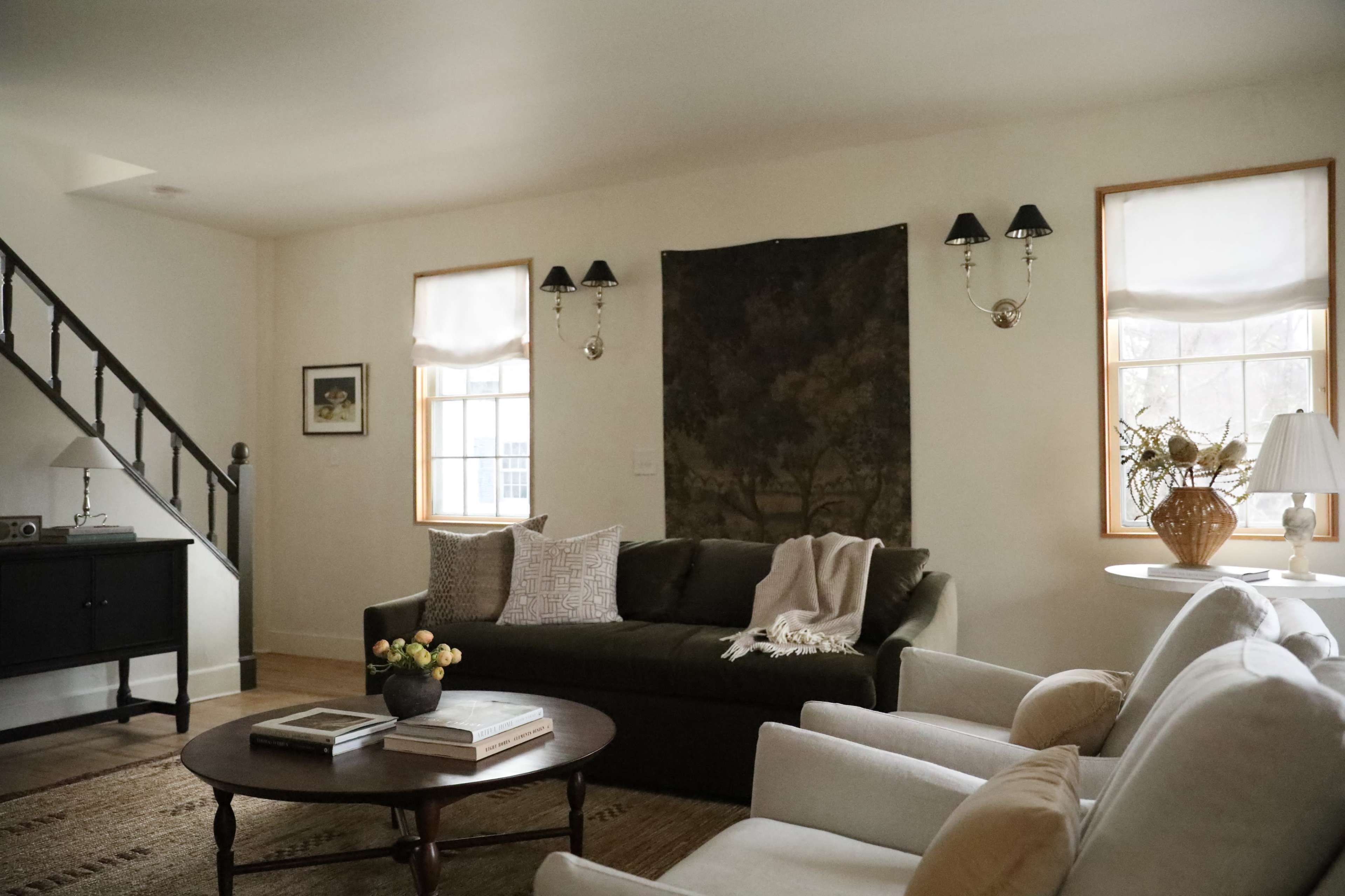 The image shows a cozy living room featuring a dark green sofa, two white armchairs, a round coffee table, and wall sconces on either side of a tapestry.