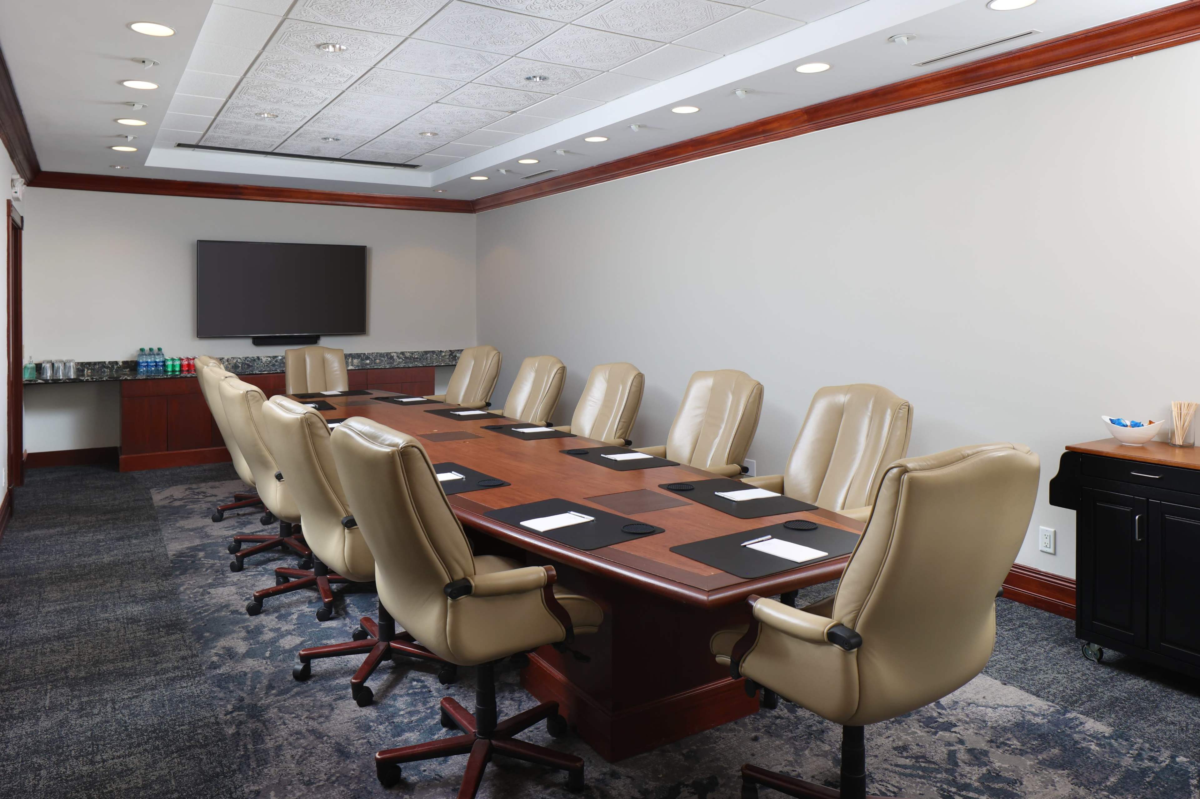 A large conference room features a long wooden table surrounded by beige leather chairs and a wall-mounted screen.