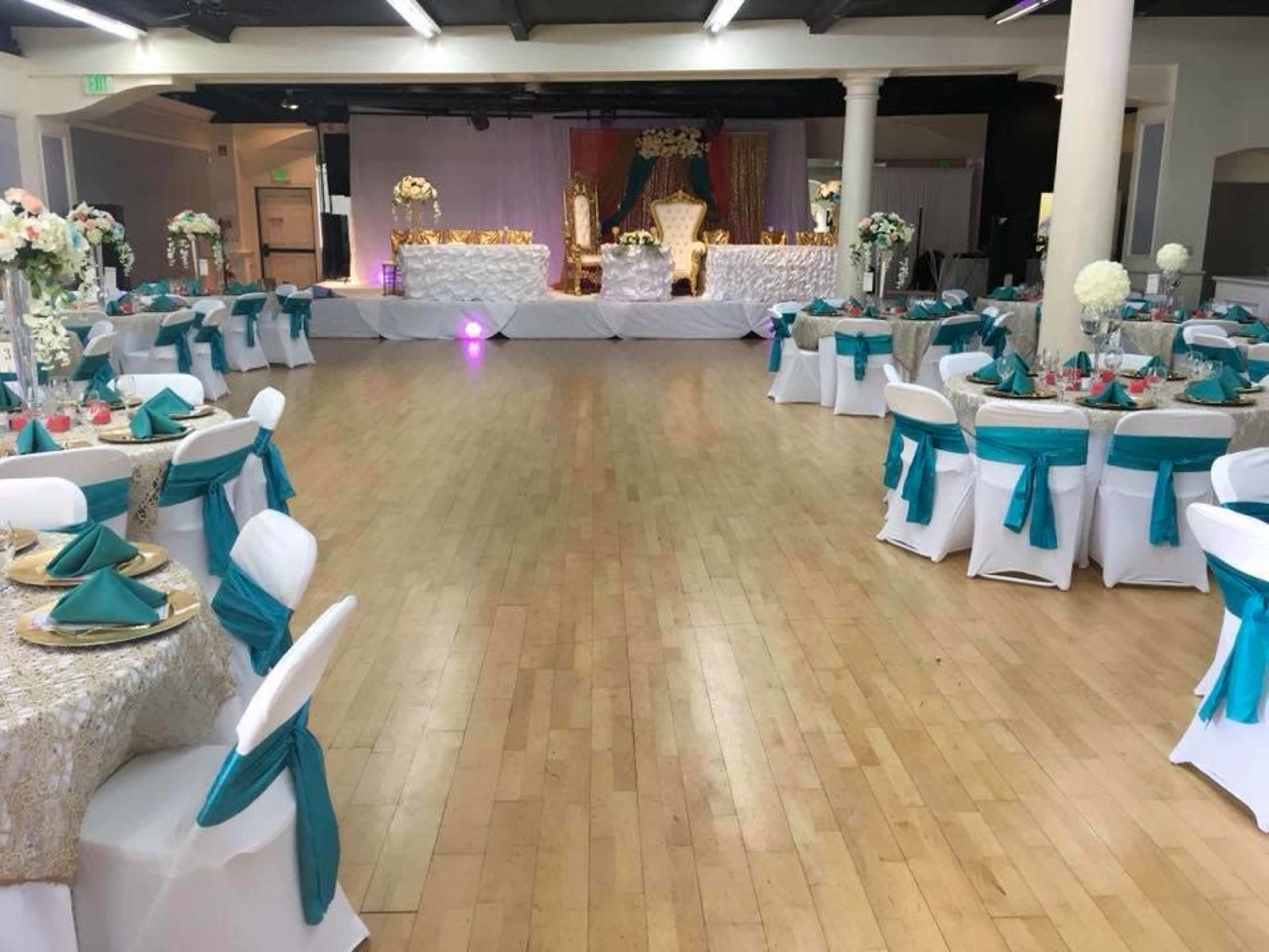 A large event space is set up with round tables adorned with white tablecloths and turquoise chair sashes, featuring floral centerpieces and a decorative backdrop at the front.
