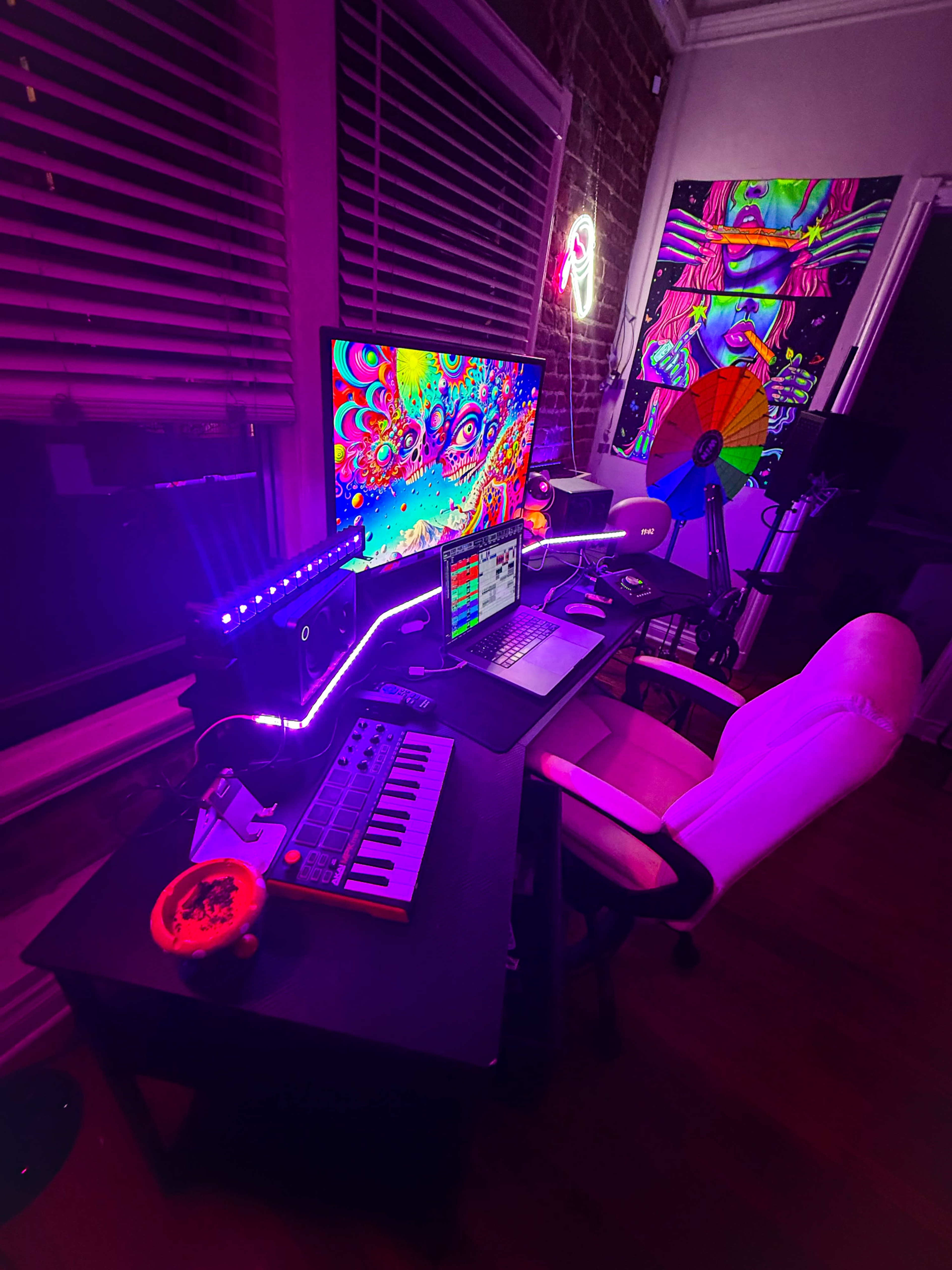 A brightly lit music production setup features a computer monitor displaying vibrant artwork, a MIDI keyboard, and a white office chair in a dimly lit room with brick walls.