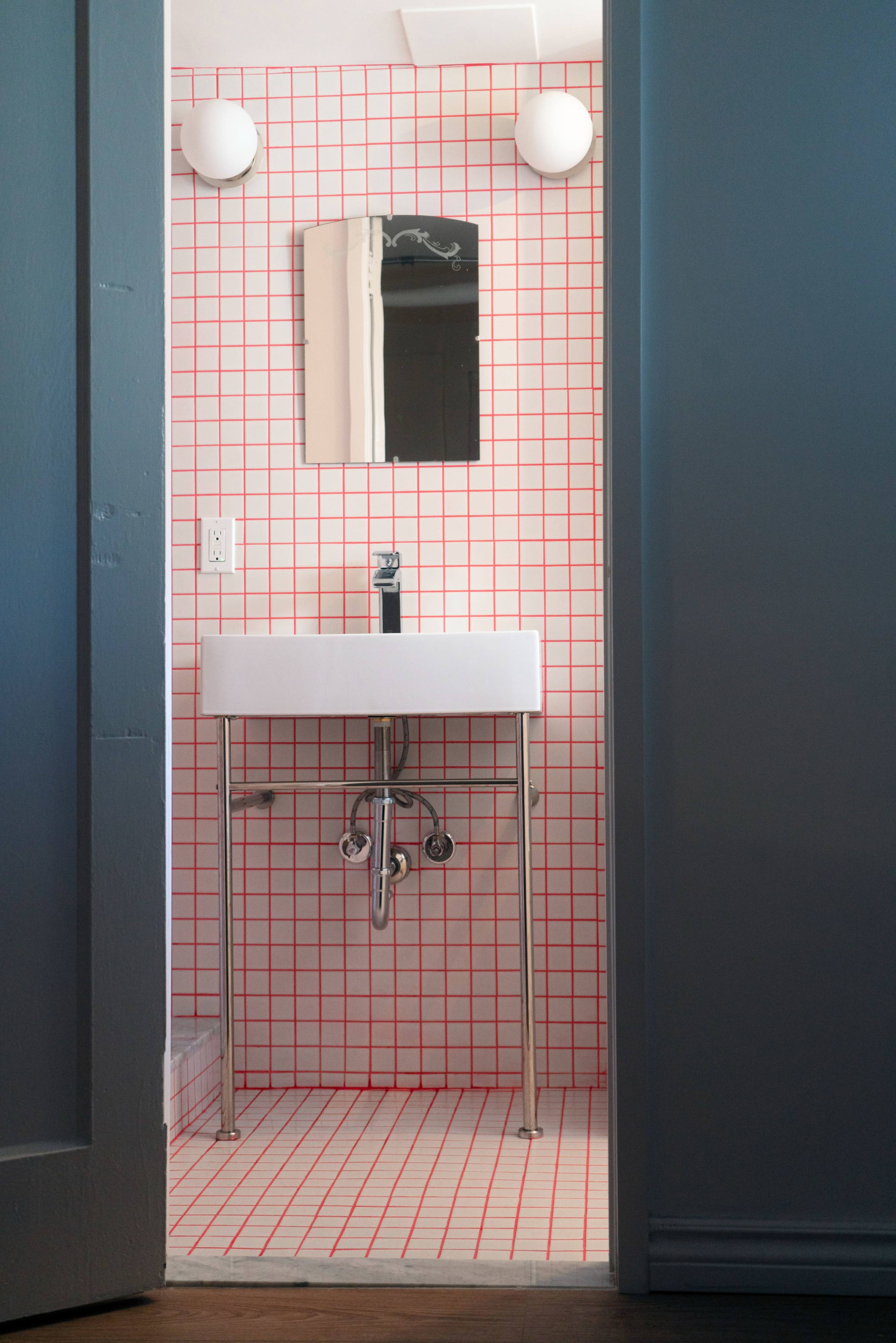 A small bathroom features a white pedestal sink against a grid-patterned wall, with a mirror and two round lights above.