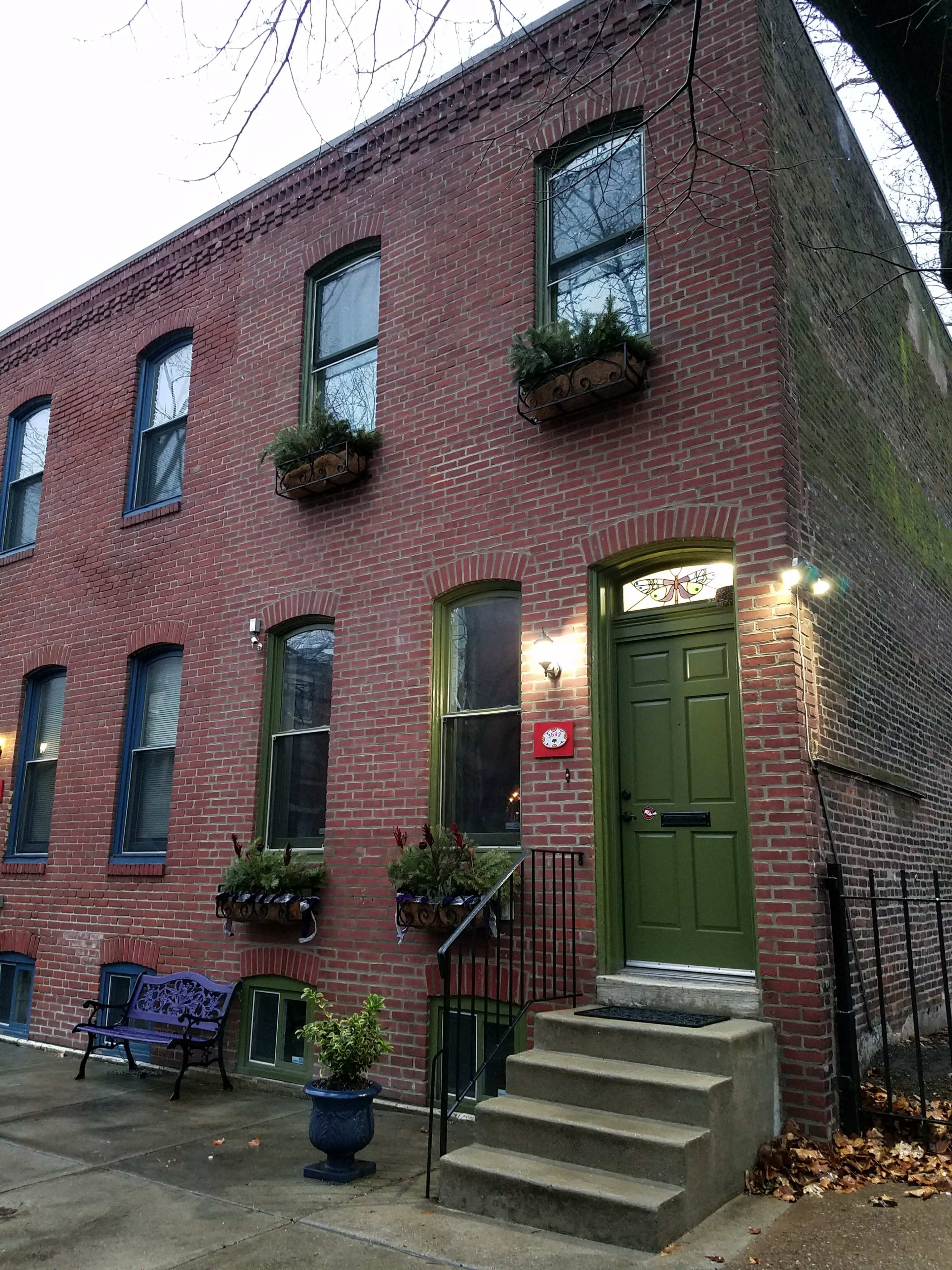 A brick townhouse with green door and window frames, featuring potted plants on the windowsills and a small staircase leading to the entrance.