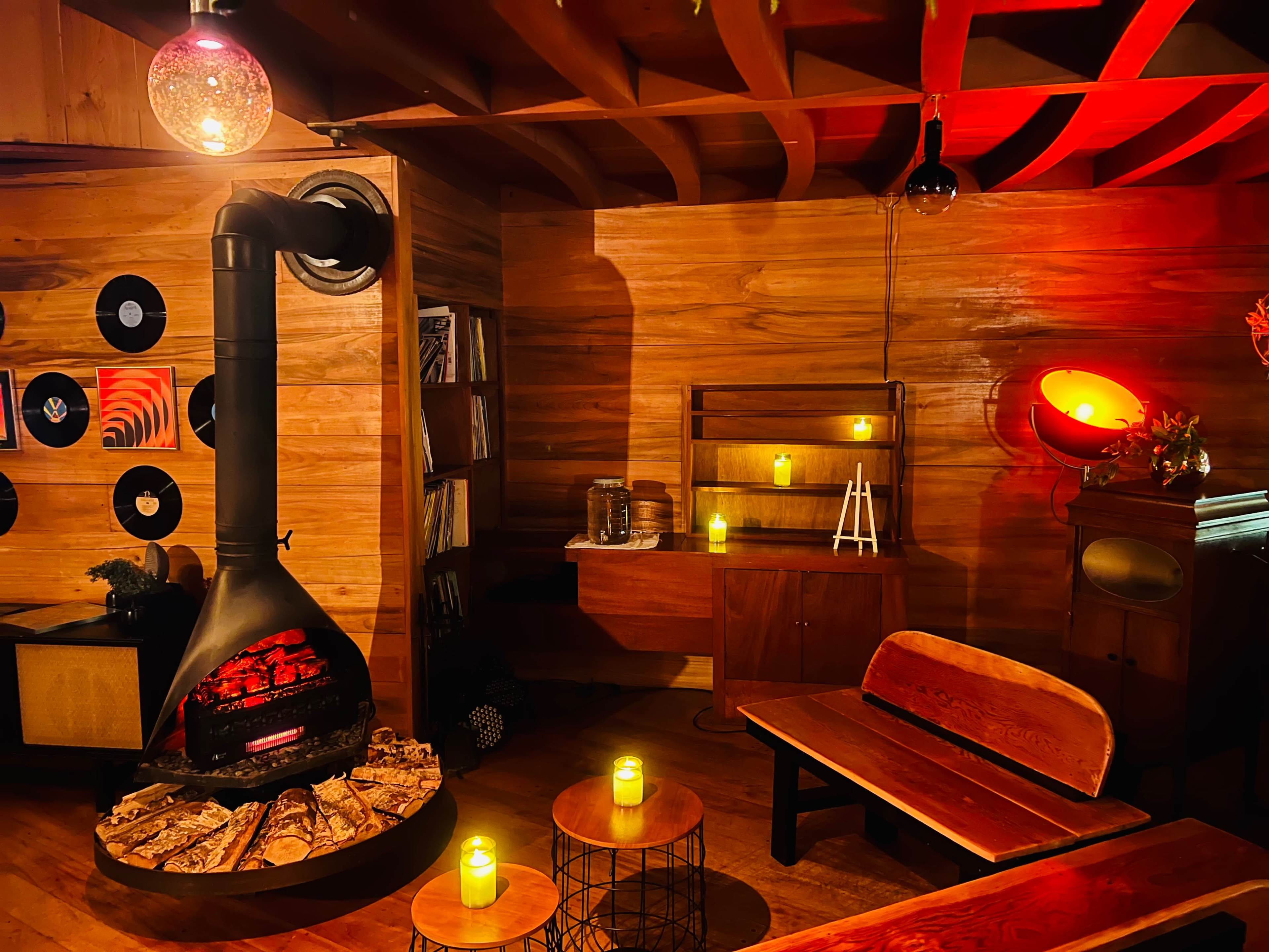 The scene features a cozy wooden room with a circular fireplace, vinyl records displayed on the walls, and candles illuminating a wooden bench and side tables.