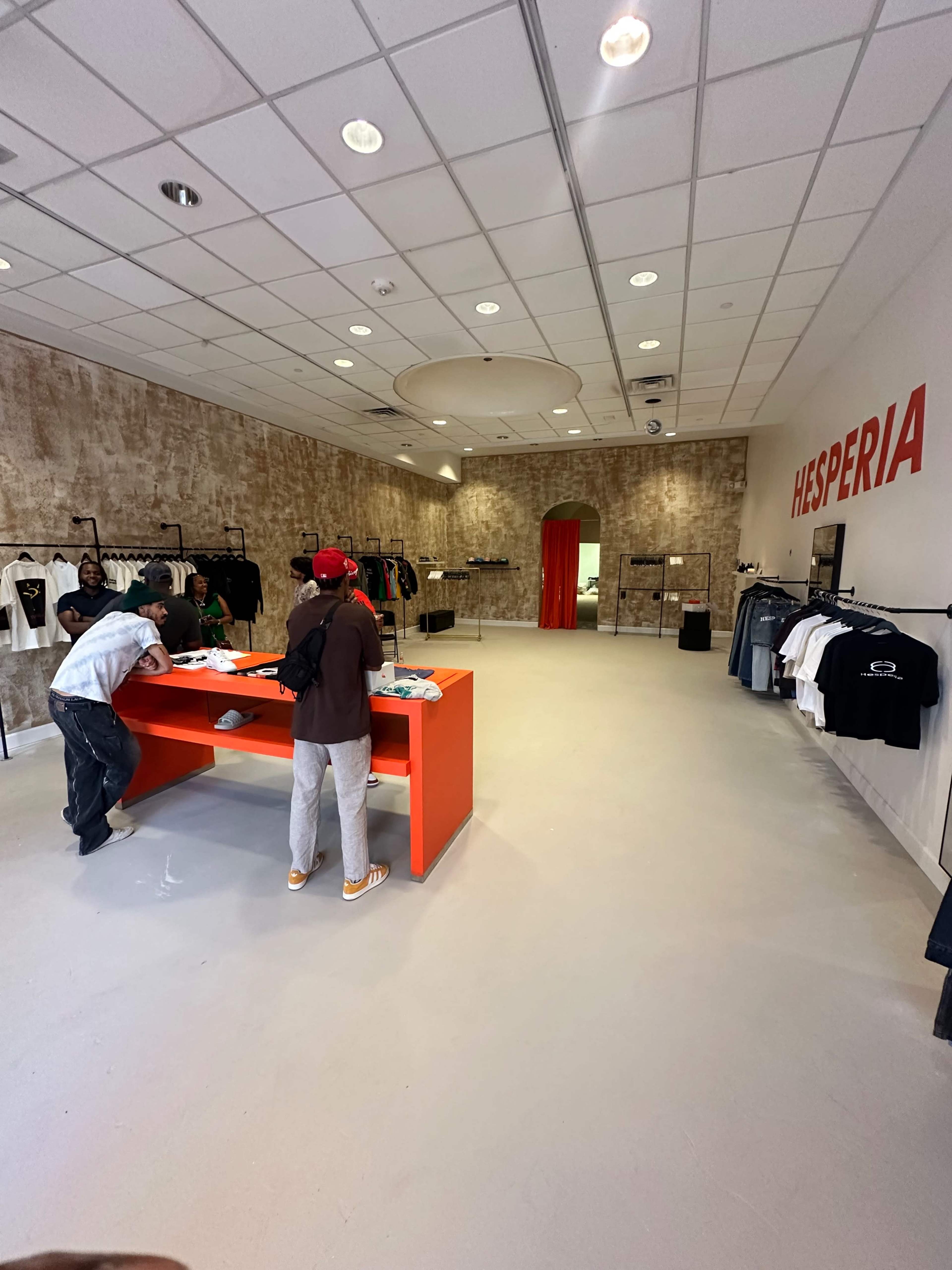 The image shows an empty clothing store with a bright orange display table in the center and racks of clothes along the walls.